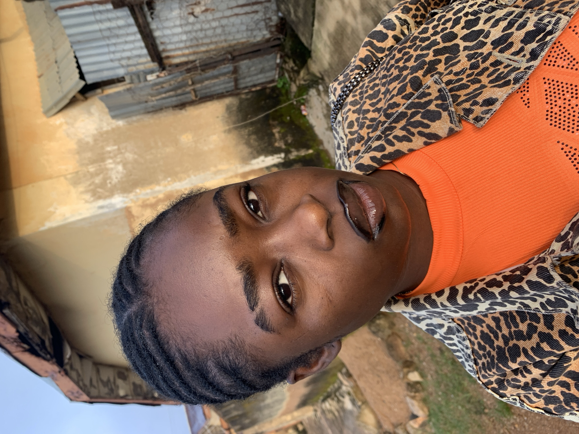 A young Black female, with braided hair and dark skin, poses for a close-up selfie outdoors. She is wearing a bright orange, ribbed long-sleeved shirt and a leopard print jacket with a black and silver chain detail. Her lips are painted with dark lipstick and a shimmering gloss. In the background, a weathered, light brown building with a corrugated metal roof and some overgrown vegetation are visible. The scene appears to be in Rafin Par, Nigeria.