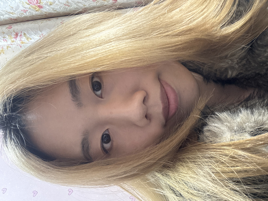 The image displays a close-up, head-and-shoulders portrait of a single individual, identified as a young East Asian female. She is positioned centrally, facing forward, and looking directly into the camera with a slight, closed-mouth smile. Her hair is long, light blonde, with prominent dark roots visible at the scalp, and it frames her face. She has dark eyes. The individual is wearing a garment with a distinct, multi-toned fur or faux-fur collar or lapel, exhibiting shades of brown, grey, and cream. The immediate background on the left consists of a light-colored, possibly white or pale pink, wall with faint, light pink heart-shaped decals. On the right, a light-colored curtain featuring a floral pattern with pink and green motifs is visible. The scene is set indoors. The specific location within Shapingba District, China, is not visually discernable from the elements present in the image.