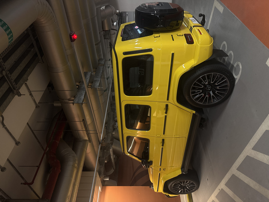 A bright yellow Mercedes-Benz G-Wagen (G-Class SUV) is stationary, parked diagonally in a designated bay within a modern, indoor parking garage. The vehicle features black multi-spoke alloy wheels with red brake calipers visible, black running boards, and "V8 BITURBO" badging on the front fender. A black spare tire cover is mounted on the rear door, and a tow hitch receiver is present below the rear bumper. The interior visible through the windows shows light brown or tan upholstery. The concrete floor has white painted parking lines and an adjacent orange-painted section. A yellow parking barrier is positioned at the front of the vehicle. The overhead exposed ceiling contains numerous large, insulated silver pipes, a prominent red pipe, electrical conduits, and structural bracing. A small red light is attached to the ceiling infrastructure above the vehicle's rear. A wall with an orange-brown gradient and subtle wave patterns is visible to the left of the vehicle. The overall environment suggests a well-maintained, contemporary parking facility consistent with those found in Dubai, United Arab Emirates, though no specific landmark is identifiable. No individuals are present.