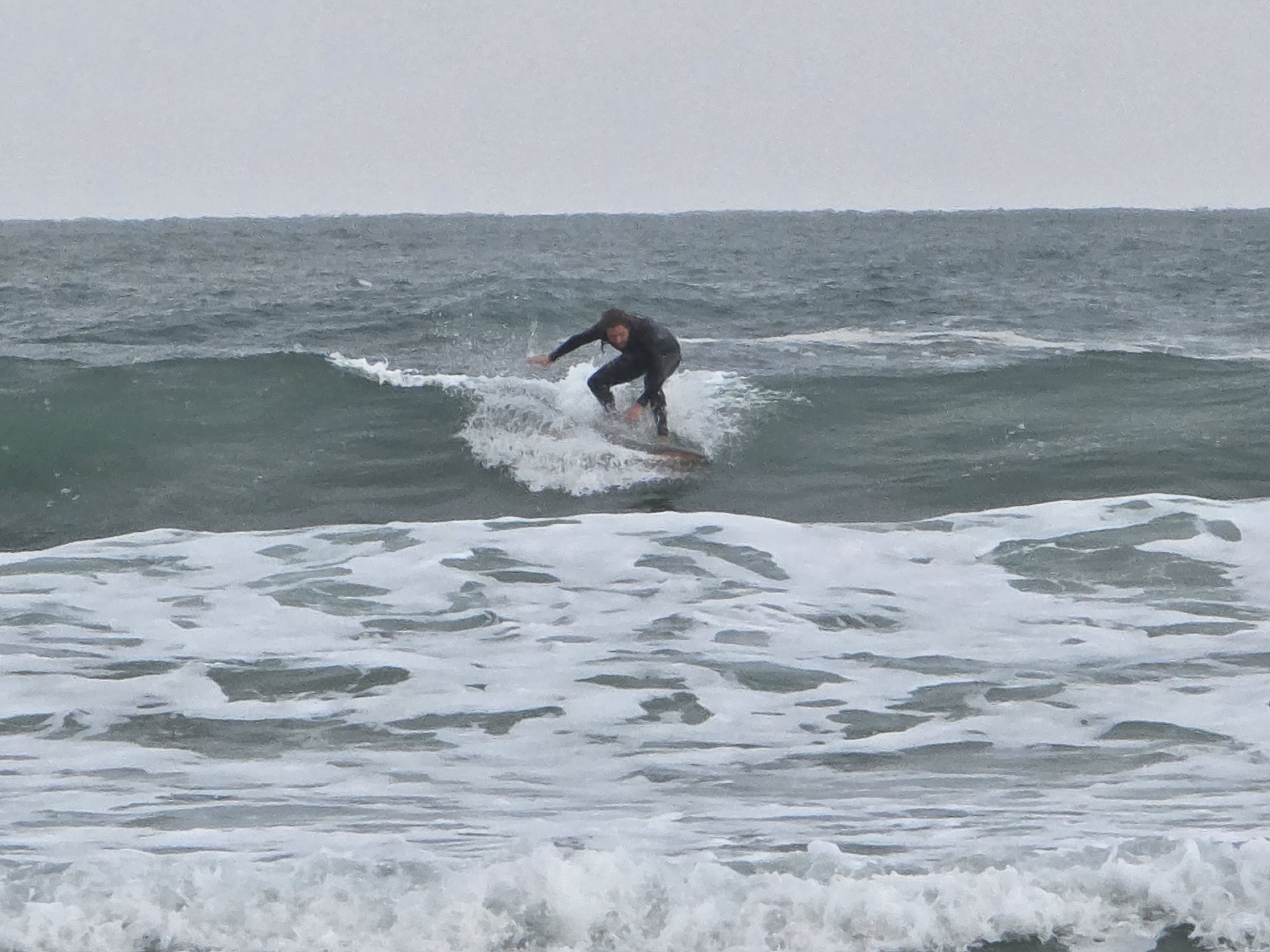 A surfer rides a wave on a cloudy day. The ocean is choppy, with white foamy waves in the foreground and larger, darker waves further back. The surfer is wearing a dark wetsuit and is crouched on a surfboard, with one arm outstretched. The sky is overcast and gray. The location is not specified as Tel-Aviv in the image or description.