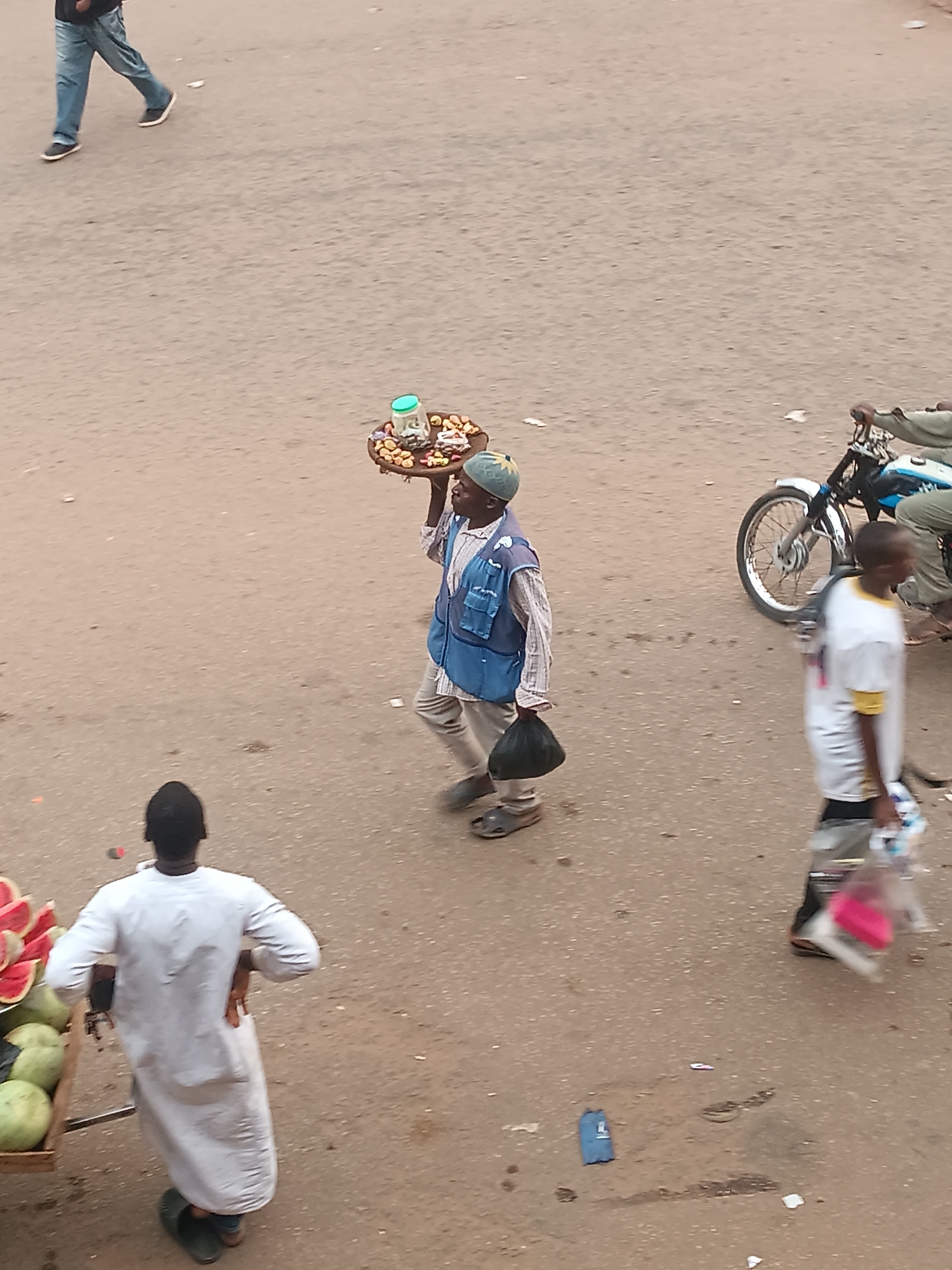 The image, likely taken from an elevated position, shows a street scene in Bukuru, Nigeria. The street appears to be paved and somewhat worn. In the foreground, a man in a white robe stands beside a cart loaded with watermelons.
A man wearing a blue cap, a striped shirt, and a blue vest carries a tray on his head. The tray holds various items, possibly food or snacks, including what appears to be a jar. He holds a black bag in his other hand.
Further away, a man in a white t-shirt and grey shorts walks with a bag in his hand. A motorcycle with two riders passes by.
In the upper left corner, a person is partially visible, wearing jeans and walking. The overall lighting suggests it is daytime.