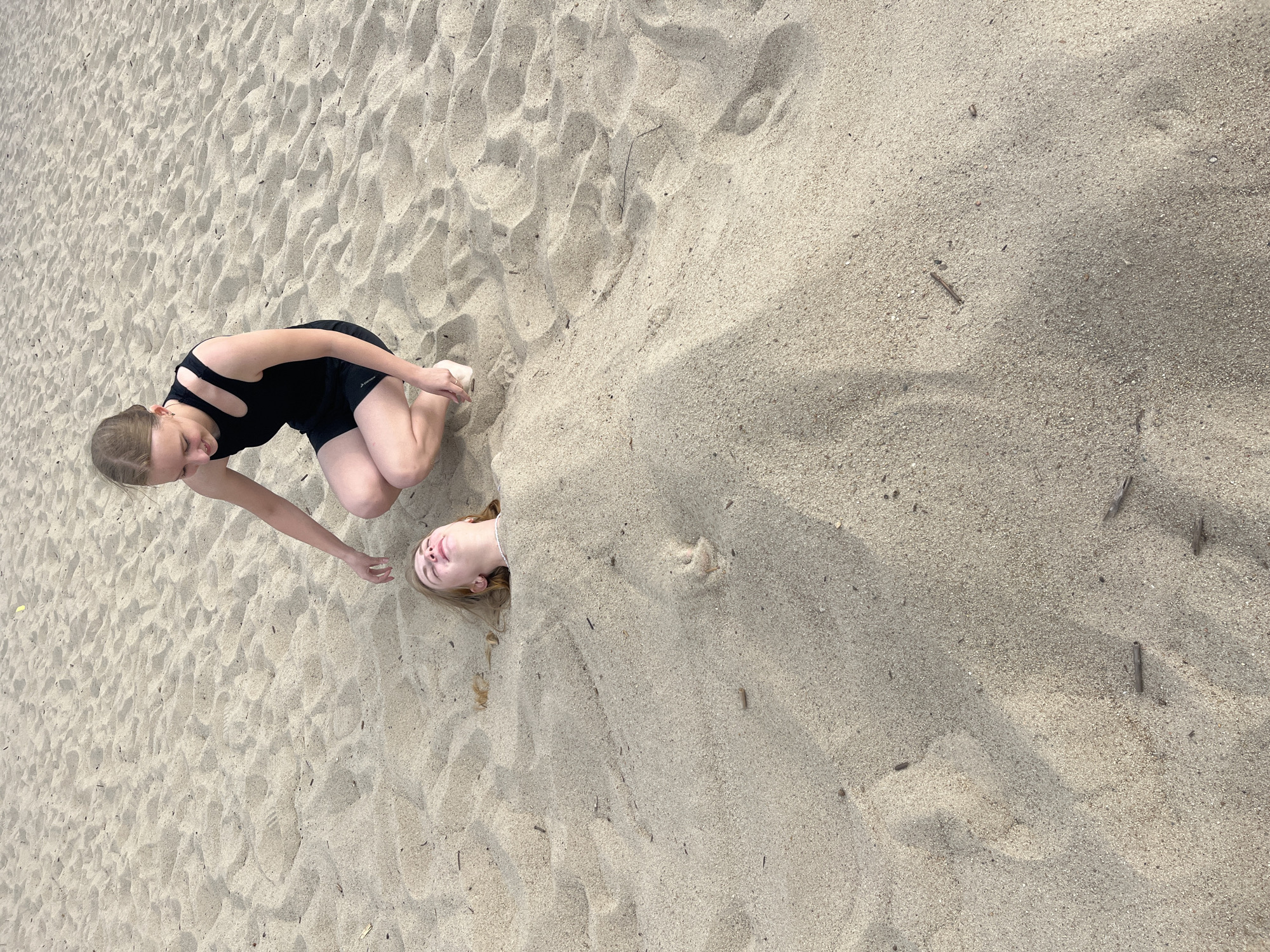 Two young women are buried in sand on a beach. The woman on the left is wearing a black swimsuit and is crouched down, reaching out with her hand. The woman on the right has her head and shoulders sticking out of the sand, with her eyes closed and a relaxed expression. The sand is light brown and textured, with some small twigs and debris scattered throughout. The lighting suggests it is daytime, possibly late afternoon, with shadows indicating the sun is at an angle. The overall mood appears playful and carefree. There is no visible text in the image.