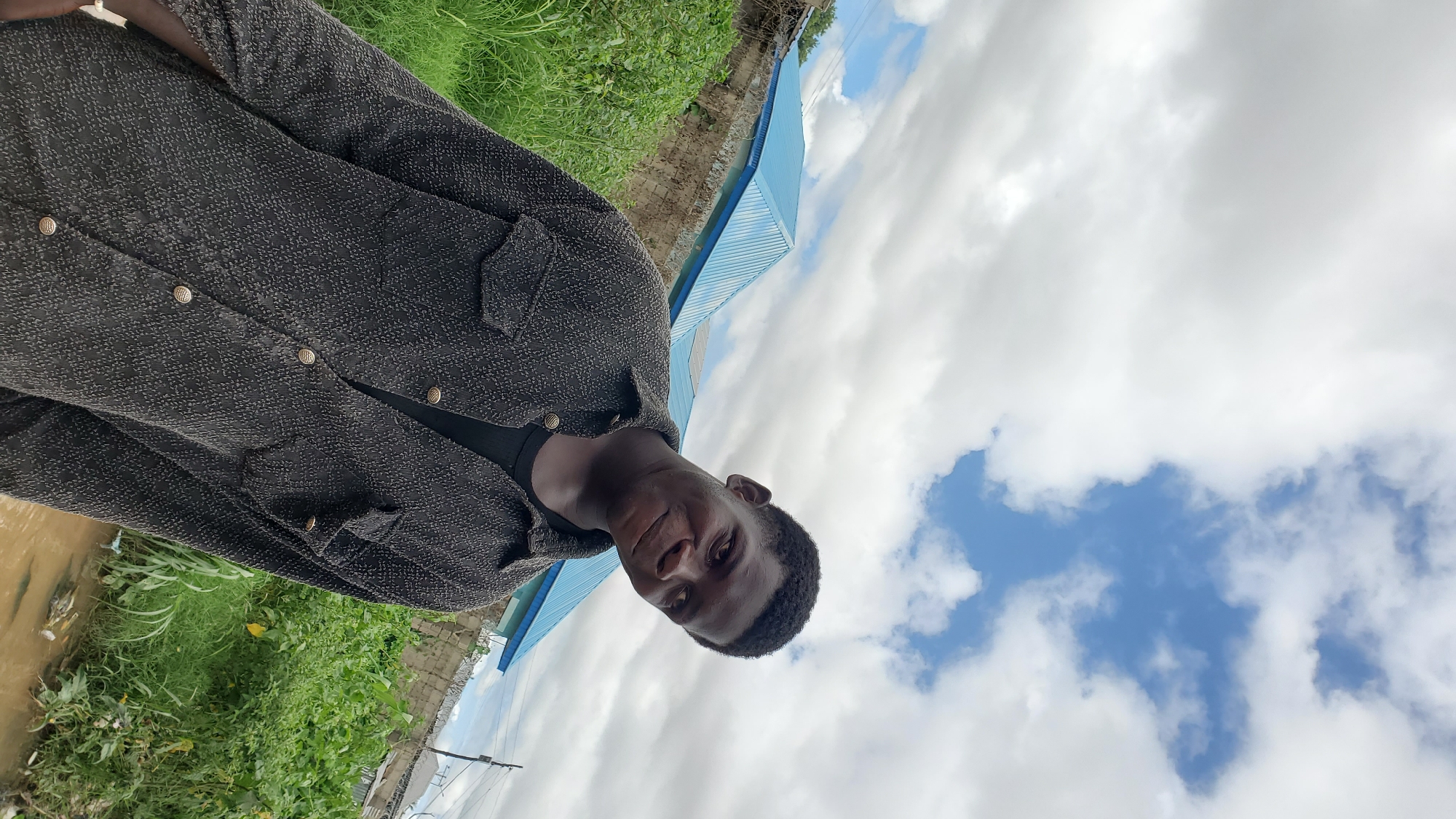 A young Black male wearing a dark textured button-up shirt over a black t-shirt is pictured outdoors. He is smiling at the camera, with his head tilted slightly. Behind him, lush green vegetation grows, and a blue-roofed building is visible, suggesting an urban or semi-urban environment, possibly in Jalingo, Nigeria. The sky above is partly cloudy. The image appears to be a casual portrait taken in a residential or undeveloped area, with some debris visible in the foreground near a water source.