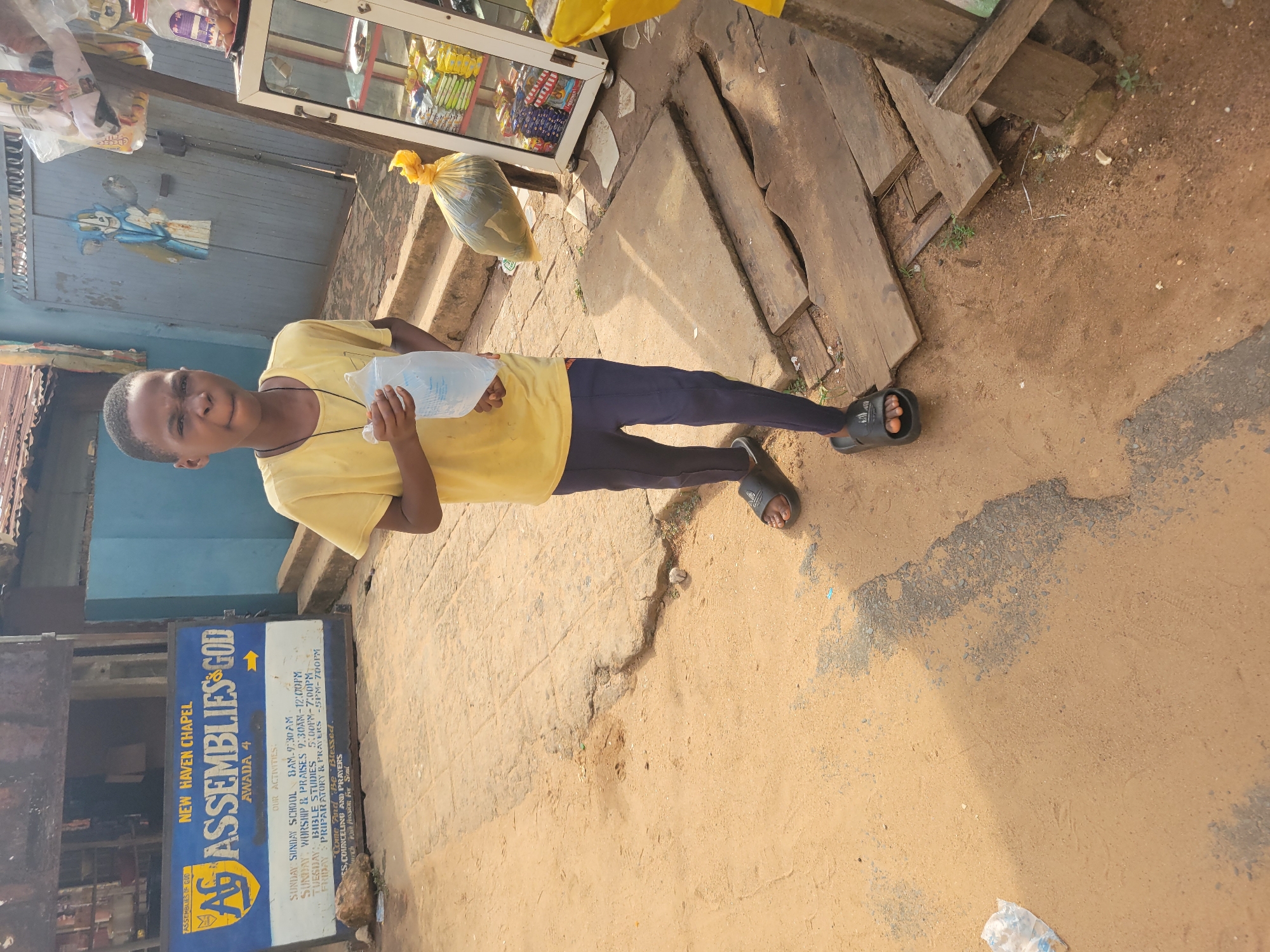 A young person stands on a dirt road in front of New Haven Chapel Assemblies of God, Awada 4, Onitsha, Nigeria. The person wears a yellow t-shirt and dark pants with black sandals, holding a water-filled plastic bag. Behind the person is a small store with various goods, and a pile of weathered wooden planks. The ground is a mix of dirt and worn pavement. A sign for the church is visible, detailing service times and activities.