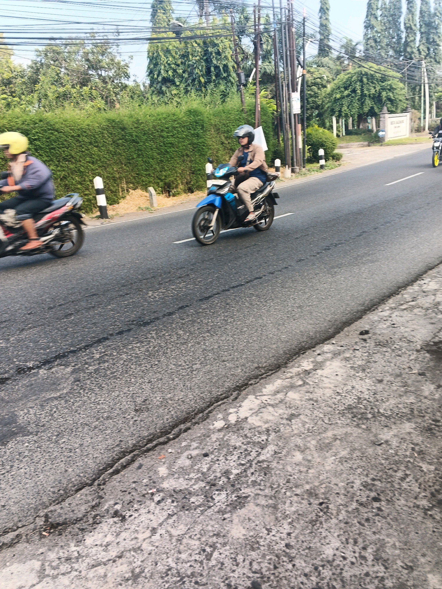 The image depicts a street scene in Ungaran, Indonesia, with a focus on two individuals riding motorcycles. In the foreground, a person wearing a yellow helmet rides a red and black motorcycle. Further down the road, another individual, wearing a dark helmet and a tan jacket over a blue shirt and beige pants, is riding a blue motorcycle. The road is asphalt, showing signs of wear with cracks and patches. To the left of the road, a dense, green hedge forms a boundary. In the background, tall trees and utility poles are visible against a clear sky, and a building with signage is partially visible on the right side of the road. The overall impression is a typical day on a road in this Indonesian city.