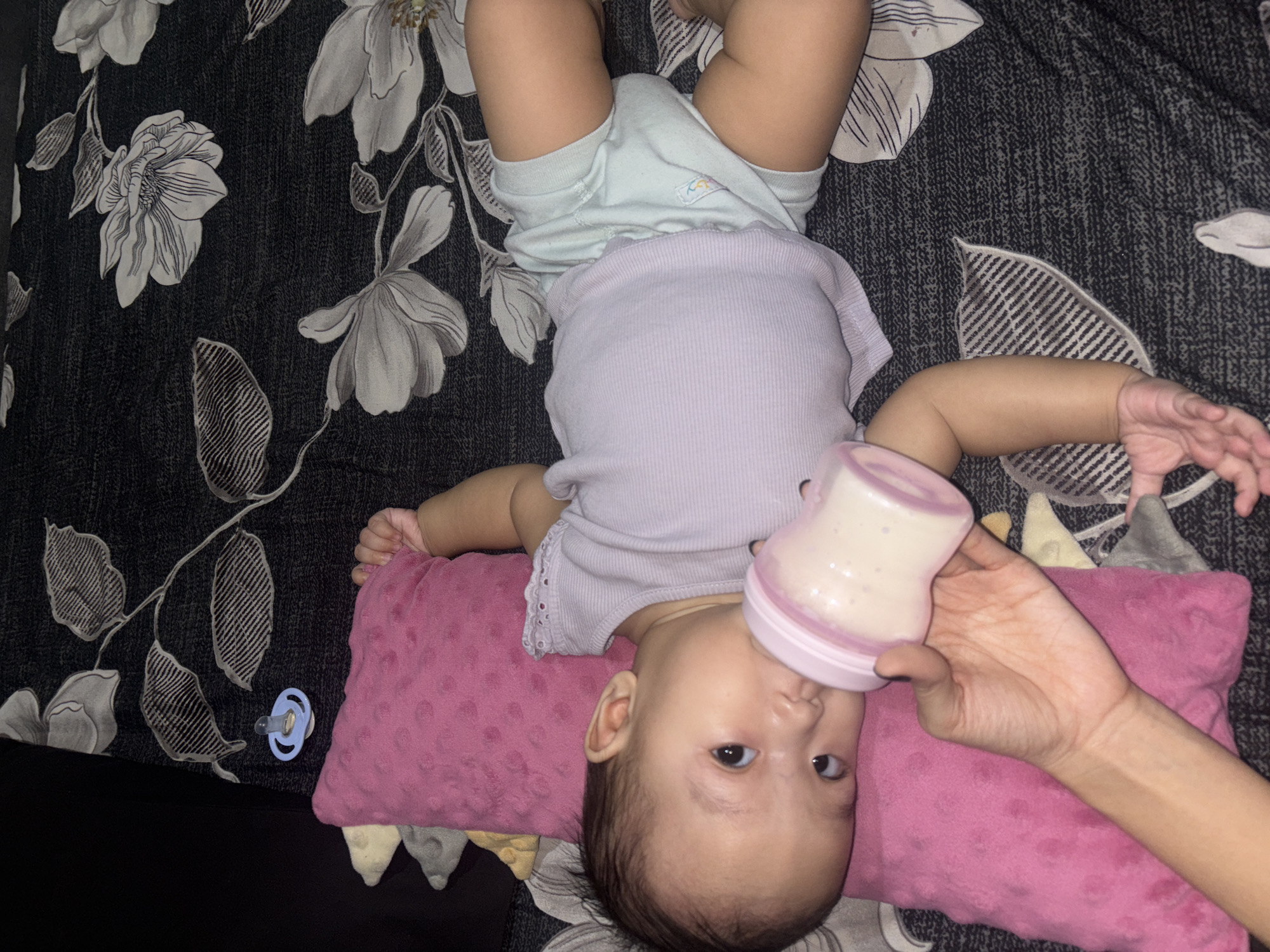 An infant is lying on its back on a patterned blanket, supported by a pink pillow. The infant is wearing a light purple ribbed tank top and light blue shorts. A hand is holding a baby bottle, offering it to the infant who is drinking from it. A pacifier lies on the blanket to the upper left of the infant. The background is a dark, floral-patterned blanket. The scene appears to be an indoor domestic setting.