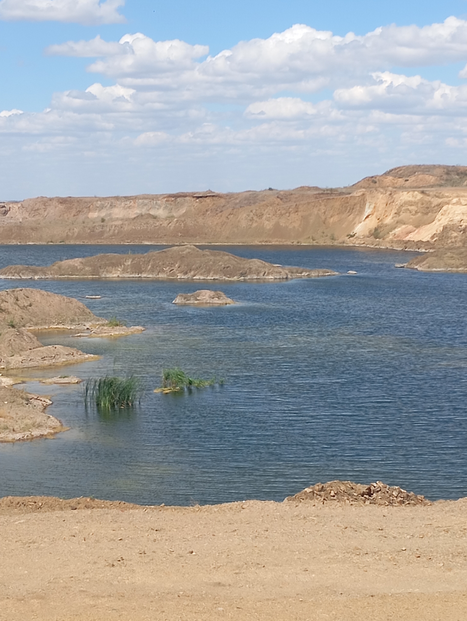 This is a vertical, eye-level, outdoor, full shot of a body of water with sandy banks, under a partly cloudy sky. The water is a deep blue, with light reflections on the surface. The banks are composed of sand and dirt, with some sparse vegetation in places. There are several small islands or peninsulas of sand and dirt jutting out into the water. In the foreground, there is a sandy embankment. In the background, there are larger, sandy hills with steep, eroded sides, suggesting a quarry or mining operation. The sky is a bright blue with scattered, puffy white clouds.