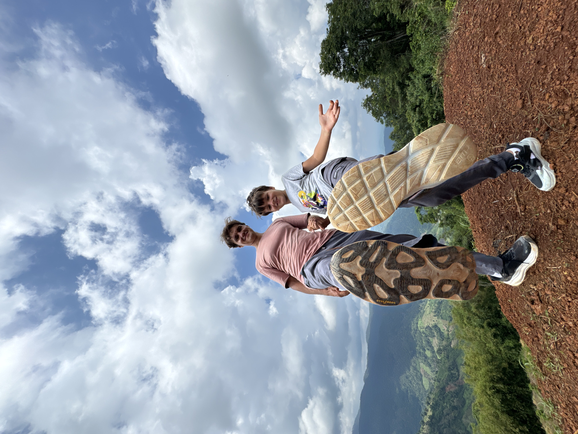 In Nao Se, Vietnam, two individuals, an adult male and a juvenile male, stand on a reddish-brown, uneven dirt surface. The adult is on the left, wearing a pink t-shirt and grey pants. His left foot is raised and angled towards the camera, showing the sole of a dark-colored sneaker with a prominent tread pattern and the "GOODYEAR" brand visible. The juvenile is on the right, wearing a grey t-shirt with a graphic print and dark pants. His right foot is raised and angled towards the camera, displaying the sole of a light-colored sneaker with a textured pattern. Both individuals are looking towards the camera with smiles. The background features a lush, green, hilly landscape under a partly cloudy blue sky. The terrain appears to be a mountainside with dense vegetation and terraced slopes visible in the distance. The perspective is from a low angle, emphasizing the sky and the raised feet of the individuals.