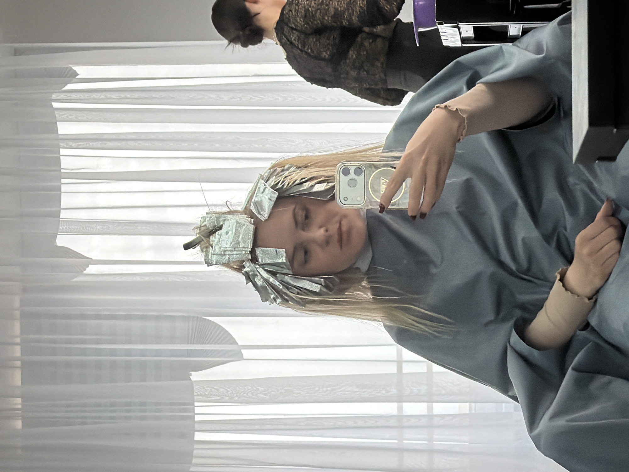 A fair-skinned female is taking a selfie while at a hair salon, as indicated by the aluminum foil in her hair. She is wearing a pale blue gown typically used in salons, and is holding a smartphone. Another woman is partially visible in the background. The setting appears to be a salon, with window blinds in the background. This image does not provide enough information to locate the specific place in the city of Votkinsk, Russia.