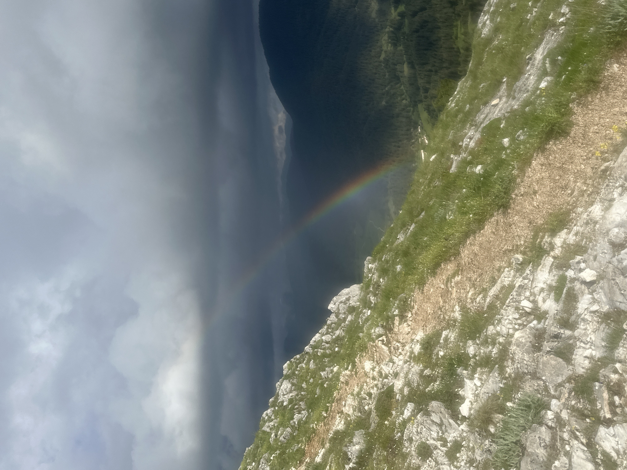 The image presents a landscape view from a high vantage point, possibly a mountain peak. The foreground features a rocky terrain with patches of grass. Beyond the rocky edge, there's a forested area. In the distance, a rainbow spans across a valley or area of lower elevation, with dark, overcast skies and rain clouds dominating the left side of the frame. The photo's orientation is sideways. There is no indication of this photo being in Iagodina, Bulgaria.