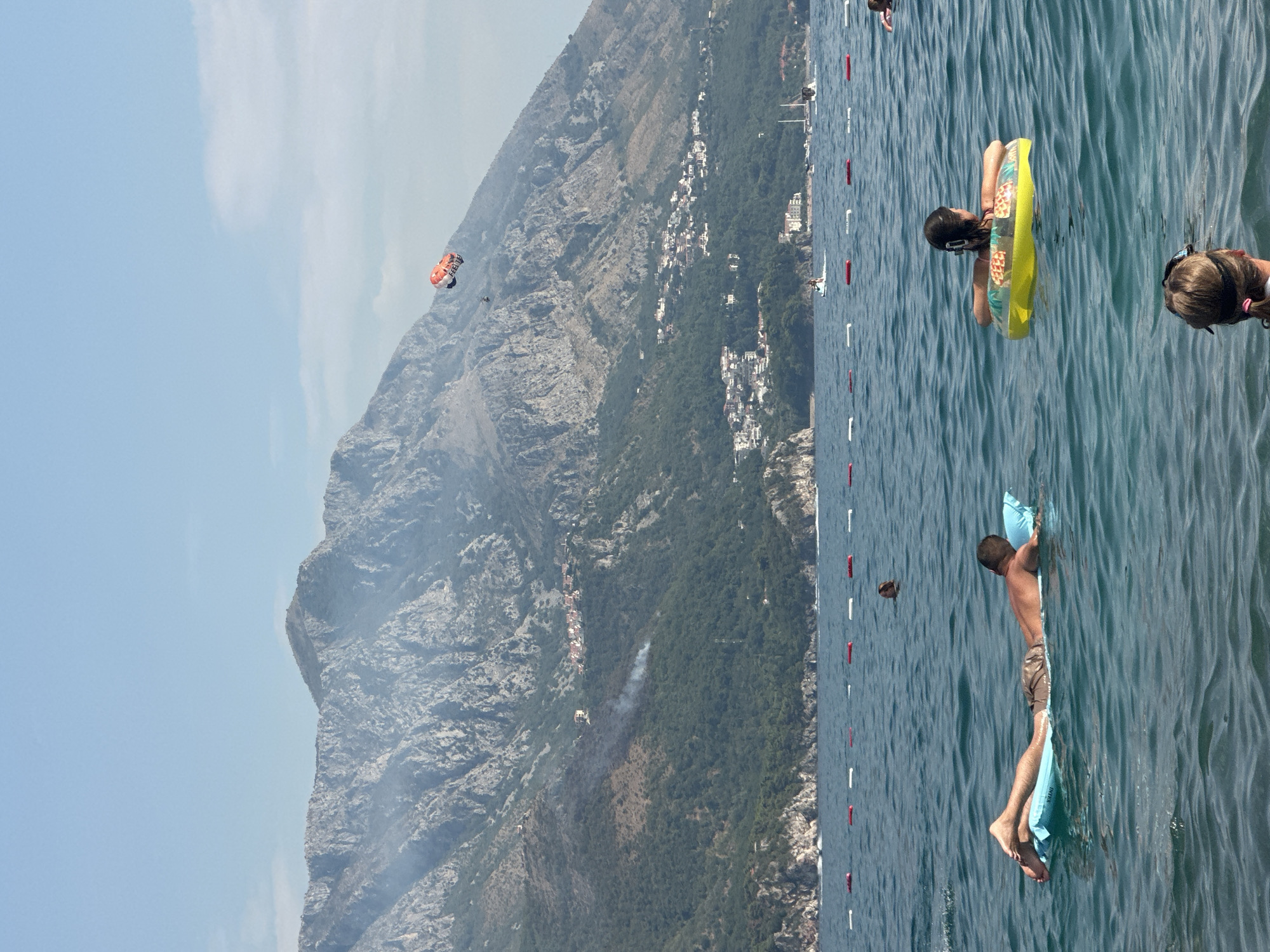 In the coastal city of Boreti, Montenegro, a tranquil seascape unfolds under a clear sky. The azure waters of the Adriatic Sea are dotted with several individuals enjoying the day. In the foreground, a man reclines on a light blue inflatable raft, his legs extended and his body partially submerged in the water. To his right, a young girl floats in a yellow inner tube adorned with a pineapple pattern, her back to the viewer. Further in the distance, another person is visible on an inflatable.
The middle ground is marked by a line of buoys, delineating a swimming area. Beyond this, the coastline is dominated by a majestic mountain range, its rugged slopes covered in lush green vegetation and dotted with small, white buildings, suggesting a settlement clinging to the hillside. A plume of smoke rises from the mountainside, hinting at a possible brush fire.
In the sky above, a parasail with an orange and white canopy drifts lazily, carrying two figures who appear to be enjoying a tandem ride. The scene is bathed in the soft light of a clear day, with a gentle breeze likely rippling the water's surface. The overall impression is one of leisure and natural beauty.