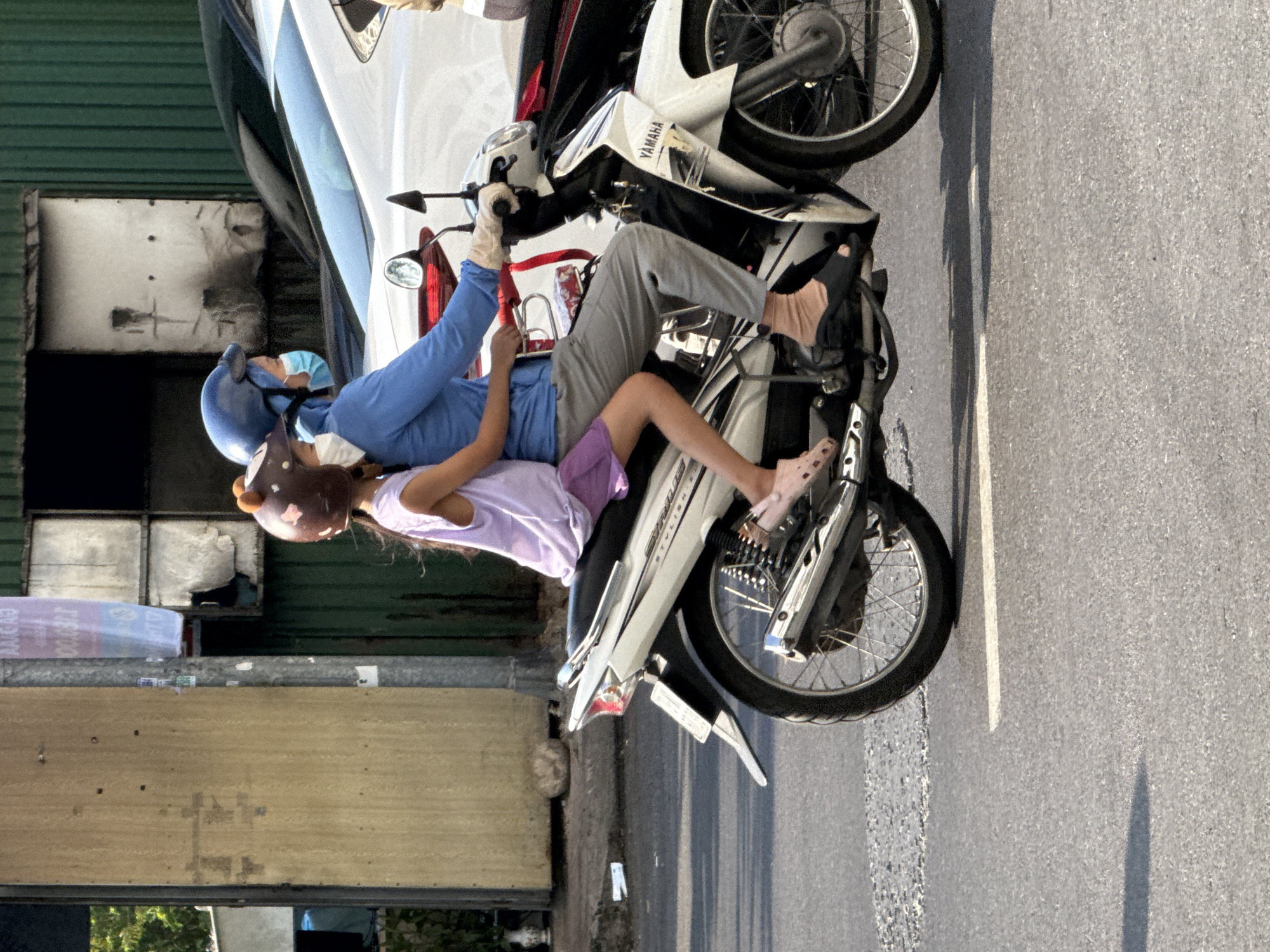 In Nam Nha Trang Ward, Vietnam, two individuals, a child and an adult, are riding a white Yamaha motorcycle on a paved road. The adult, wearing a blue long-sleeved shirt, gray pants, a blue helmet, and a light blue face mask, is operating the motorcycle. The child, in a light purple sleeveless top and a purple skirt, is seated in front of the adult, wearing a brown helmet with ear-like embellishments. Both are wearing face masks. The motorcycle has a visible "YAMAHA" logo on its front panel. In the background, a building with green corrugated metal siding and a wooden facade is visible, along with parts of other vehicles. The road surface shows signs of wear, with a white line marking a lane. Sunlight casts shadows on the road.