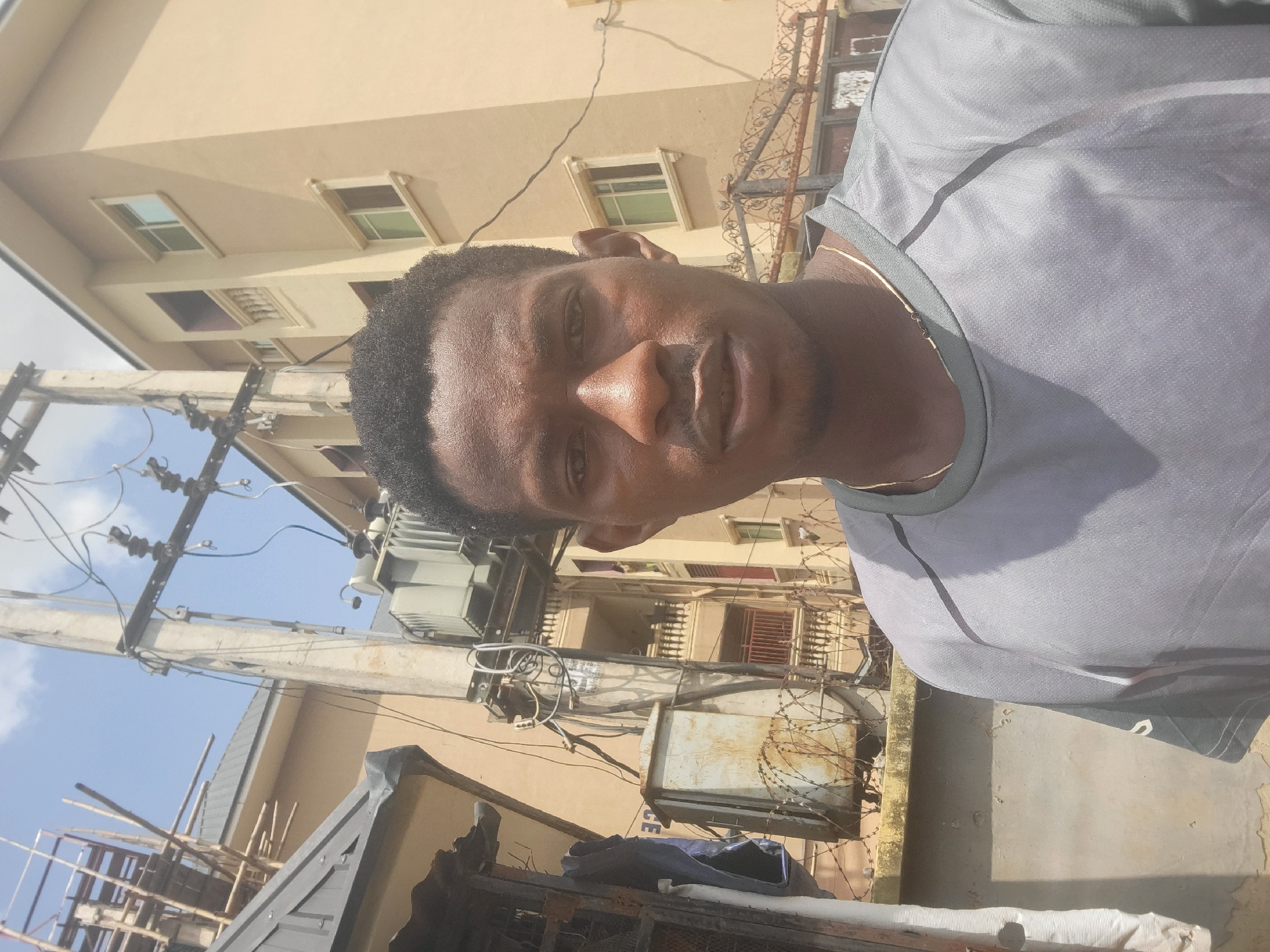 A medium shot selfie shows a dark-skinned man with a short haircut, wearing a gray t-shirt and a thin gold chain. He is standing outdoors in front of a transformer and utility pole. The background features a light brown building with several windows and balconies, and scaffolding on the left side. A rusty metal fence with barbed wire is visible in the foreground. The sky is partly cloudy and the scene appears to be in a city.