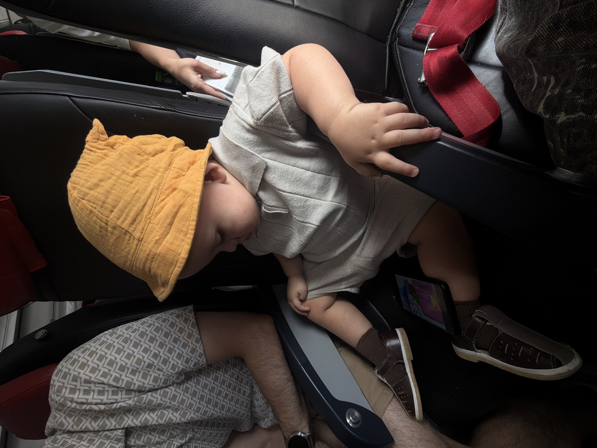 A baby or toddler is seen resting horizontally across two black leather airplane seats, presumably in a dimly lit cabin. The child wears a crinkled orange bucket hat and a light-colored, possibly beige, textured romper or shirt. Their head is turned to the left, and eyes are closed, suggesting they are asleep or resting.
In the upper left background, an adult's hand, possibly the parent's, is visible holding a white object, possibly a napkin or tissue. To the lower left, an adult's legs, wearing light-colored patterned shorts, and an arm with a smartwatch are visible, positioned below the resting child.
The child's feet, clad in brown socks and brown closed-toe sandals, rest near a dark-colored smartphone or small tablet which is displaying a colorful image or video. A red seatbelt is visible buckled into the seat in the upper right quadrant of the image. A grey and black armrest or tray table panel with a circular button separates the child from the adult in the lower left.
The setting is clearly an airplane cabin, indicated by the seat design, seatbelt, and panel features. The subdued lighting suggests it could be nighttime or a flight during which the cabin lights are dimmed. The activity is the child resting or sleeping comfortably during a flight. No discernible text is visible on any objects, including the device screen. The location context is an airplane in or around Domodedovo, Russia.