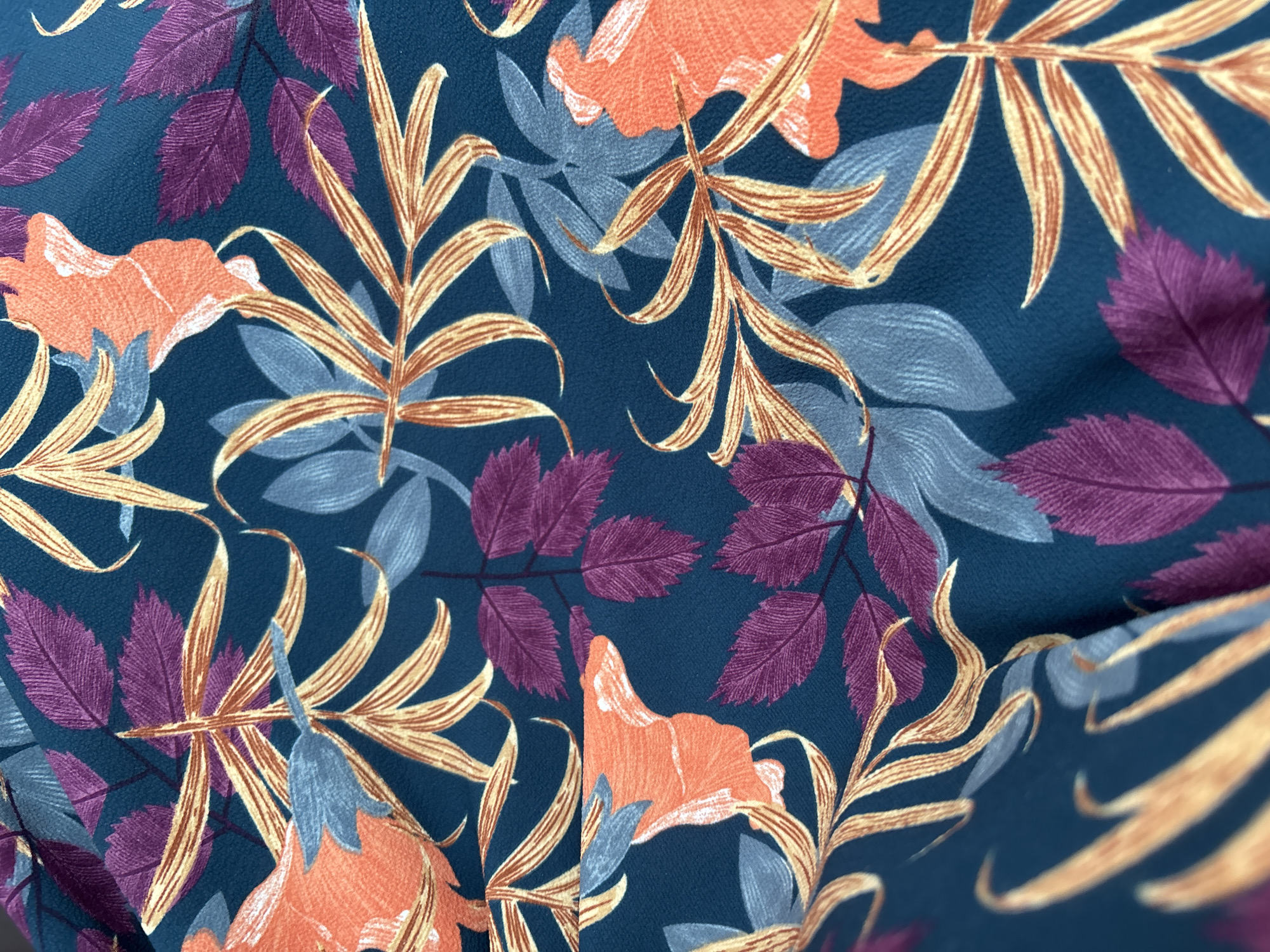 The image is a close-up, high-angle shot of a dark teal fabric with a floral and leaf pattern. The pattern features stylized elements in orange, purple, light blue, and gold. Specifically, there are large orange floral shapes that appear to be hibiscus or similar tropical flowers, intermingled with feathery gold leaves and clusters of purple and light blue leaves. The fabric is draped, creating folds and shadows that add depth and texture to the image. The lighting is even, highlighting the details of the print and the fabric's weave. There are no people or identifiable landmarks present in the image, and the scene appears to be indoors or in a studio setting, focusing solely on the textile.