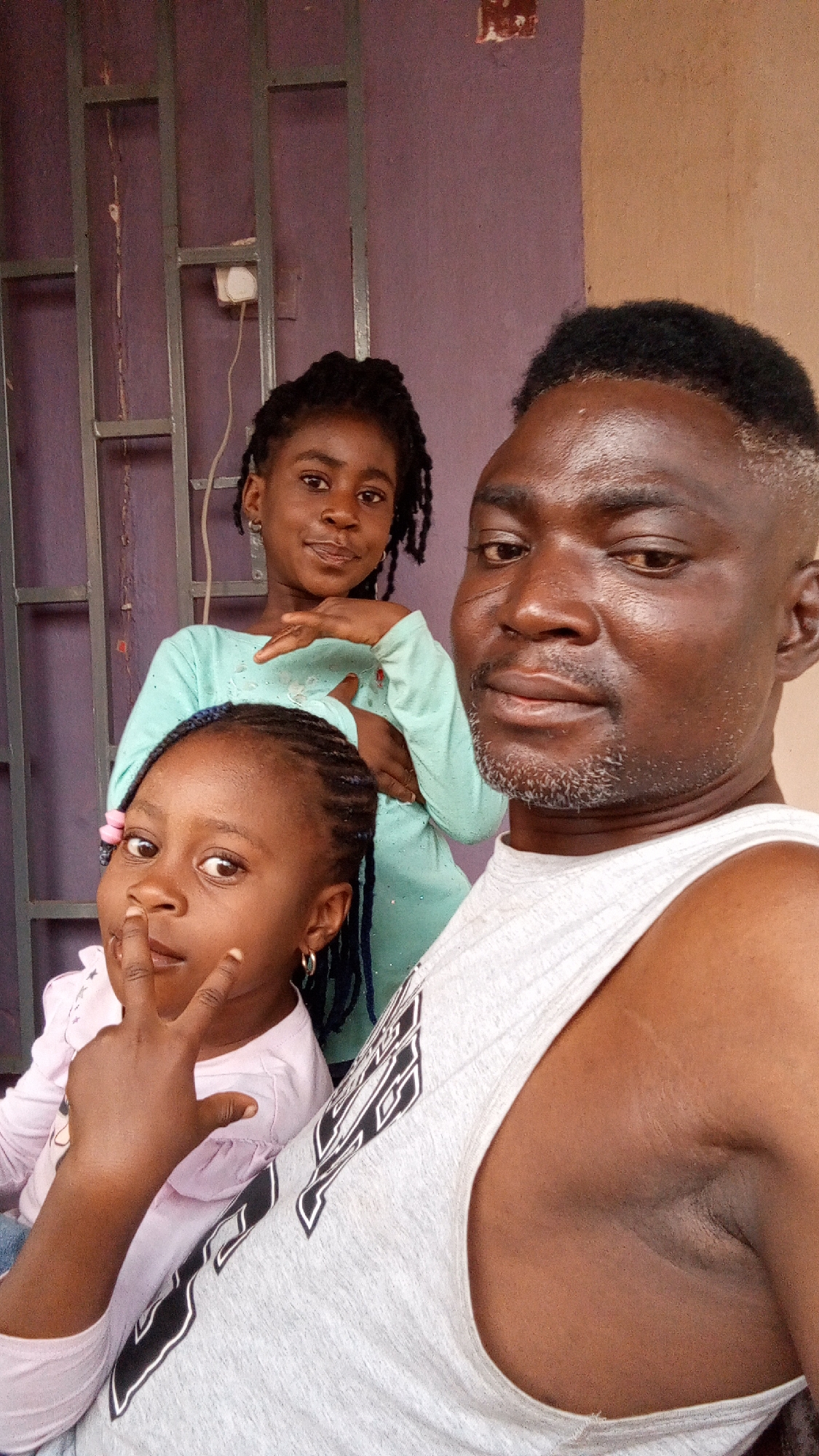 The photograph shows three people. A man is in the foreground, partially visible, wearing a white sleeveless shirt. Behind him and to his left are two young girls. The girl in the front wears a pink long-sleeved shirt and has a pink flower in her hair. She is holding up two fingers. The other girl, standing slightly behind and to the right of the first girl, wears a light green long-sleeved shirt. The background includes a purple wall with a metal grate design and a light-colored wall. The location is inferred to be Sabon Tasha, Nigeria. The image depicts a posed photograph of the three people.