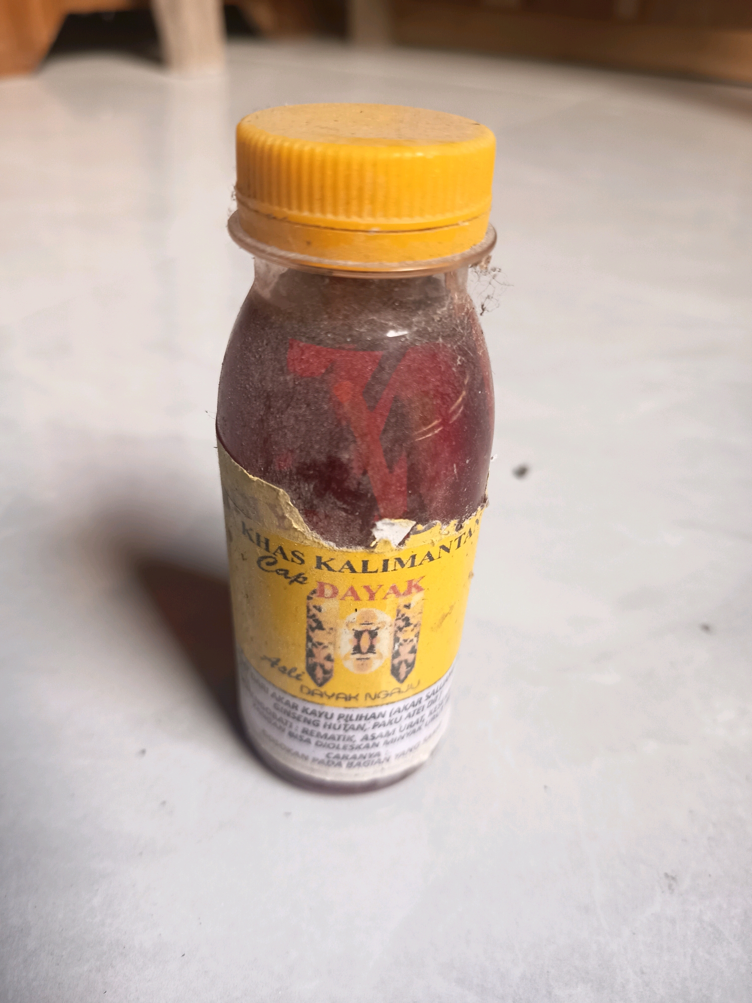 A close-up, eye-level shot shows a small glass bottle with a yellow cap, resting on a light-colored, textured surface that appears to be a floor or table. The bottle is partially filled with a dark red liquid and covered in dust. A torn yellow label wrapped around the bottle has text in both English and what appears to be Indonesian. The visible text on the label includes "KHAS KALIMANTAN," "Cap DAYAK," and "ASLI DAYAK NGAMBAU." Below this, smaller text is partially obscured by dust and damage but seems to describe ingredients and usage. The background is out of focus, showing hints of wooden furniture in a dimly lit indoor environment, potentially in a residential area of Sidorejo, Indonesia. The lighting is dim and appears to be artificial, casting soft shadows.