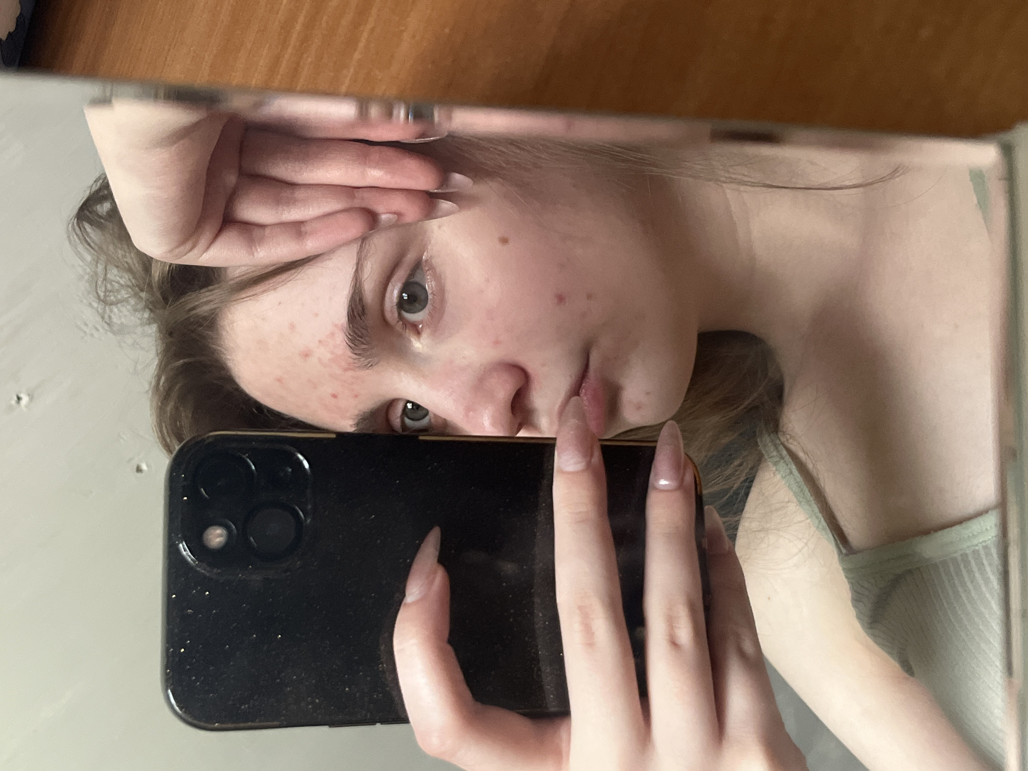 The image shows a young woman in a close-up, mirror selfie. She is holding a black smartphone in her left hand, partially obscuring her face. Her right hand is resting on her forehead. She has light skin with some acne. Her hair is brown and slightly messy. She is wearing a light green ribbed tank top. The background appears to be a bathroom with a light-colored wall and a wooden surface on the right side of the frame, likely part of a vanity or cabinet. The lighting is soft. There is no indication of the city of Ashkadar, Russia in the image.