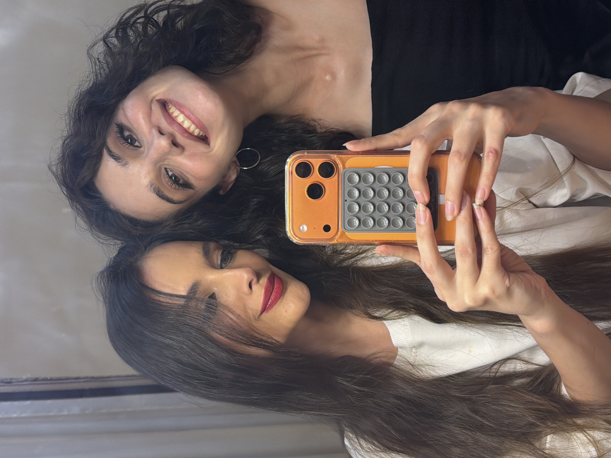 The media file is a vertically oriented selfie taken by two women, appearing to be in an indoor setting, possibly a bedroom given the soft lighting and what looks like a bed or couch with white bedding. The woman in the foreground, closer to the camera, is looking directly at the lens with a slight smile and red lipstick. Her dark, long hair is spread out around her. She is holding an orange-colored phone with a case that has a grid of circular bumps on the back. Her hand is positioned to take the picture.
Behind her, the second woman is also looking towards the camera, with a wide, genuine smile. She has dark, curly hair and is wearing a black top. A silver hoop earring is visible. The lighting in the image is soft and warm, suggesting it might be daytime, but artificial lighting could also be the source. The overall mood appears cheerful and intimate, typical of a casual selfie between friends. The reflective surface below them suggests they might be lying on a bed or floor near a mirror or other reflective surface. No text is visible in the image. The location context is Ufa, Russia.