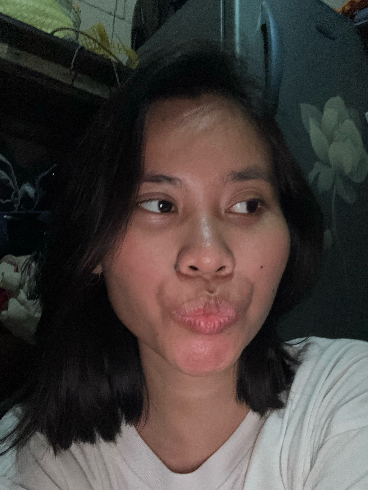 A self-portrait of a woman is captured in close-up. The woman has dark hair, a mole on her right cheek, and is puckering her lips. She is wearing a white t-shirt. The background is dimly lit and appears to be indoors, with some objects visible on shelves in the upper left corner and a patterned surface on the right. The image's lighting suggests it was taken in a low-light environment, possibly at night. The location is stated to be East Jakarta, Indonesia, though no specific landmarks or details within the image confirm this location.