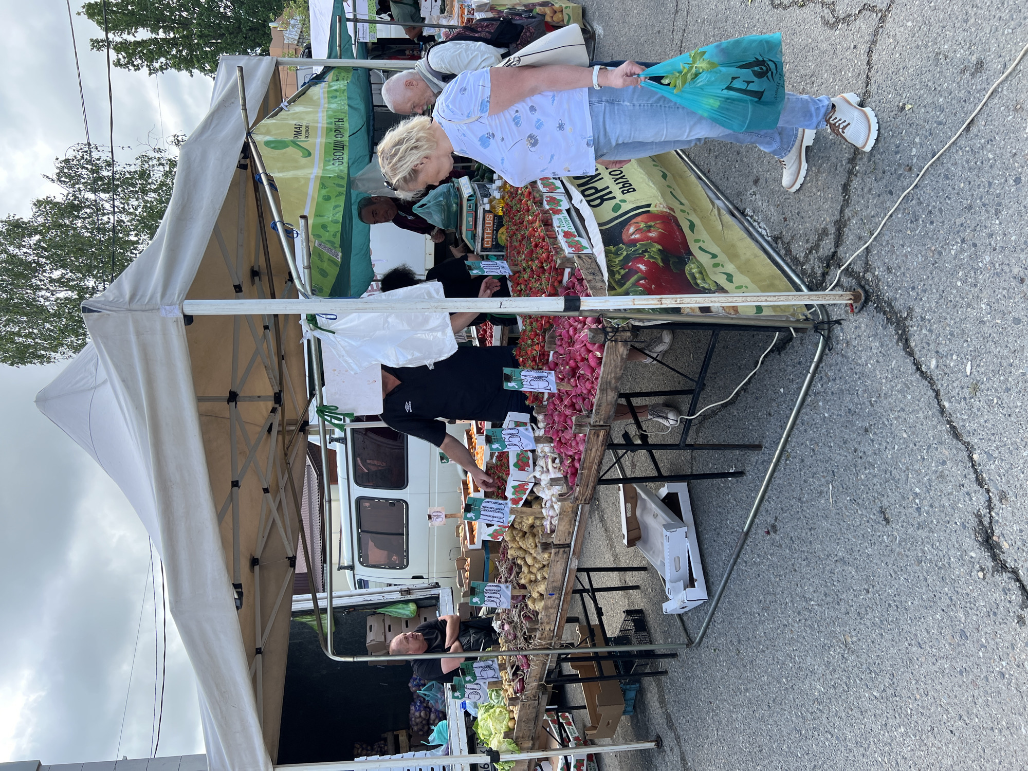 The image shows a street market in Goryachiy Klyuch, Russia. Under a white and green canopy, vendors are selling produce. A woman in a white shirt and jeans stands at a table covered with strawberries and radishes, holding a blue plastic bag. Other customers and vendors are visible. A white van is parked behind the market stall. The ground is asphalt.