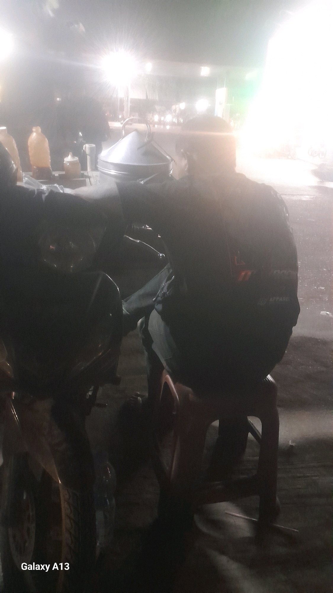 The image depicts a nighttime scene in Jekulo, Indonesia. Two individuals are present, one seated on a red plastic stool behind a motorcycle. This person is wearing dark clothing with a visible logo on the back. They appear to be interacting with a food cart, which has various containers and a metal steaming pot. Another individual is partially visible on the left, seemingly standing next to the motorcycle. The environment is dimly lit, with bright lights in the background suggesting a street or marketplace setting. The ground appears wet, indicating recent rain or damp conditions. The overall atmosphere is that of a street food stall operating at night.