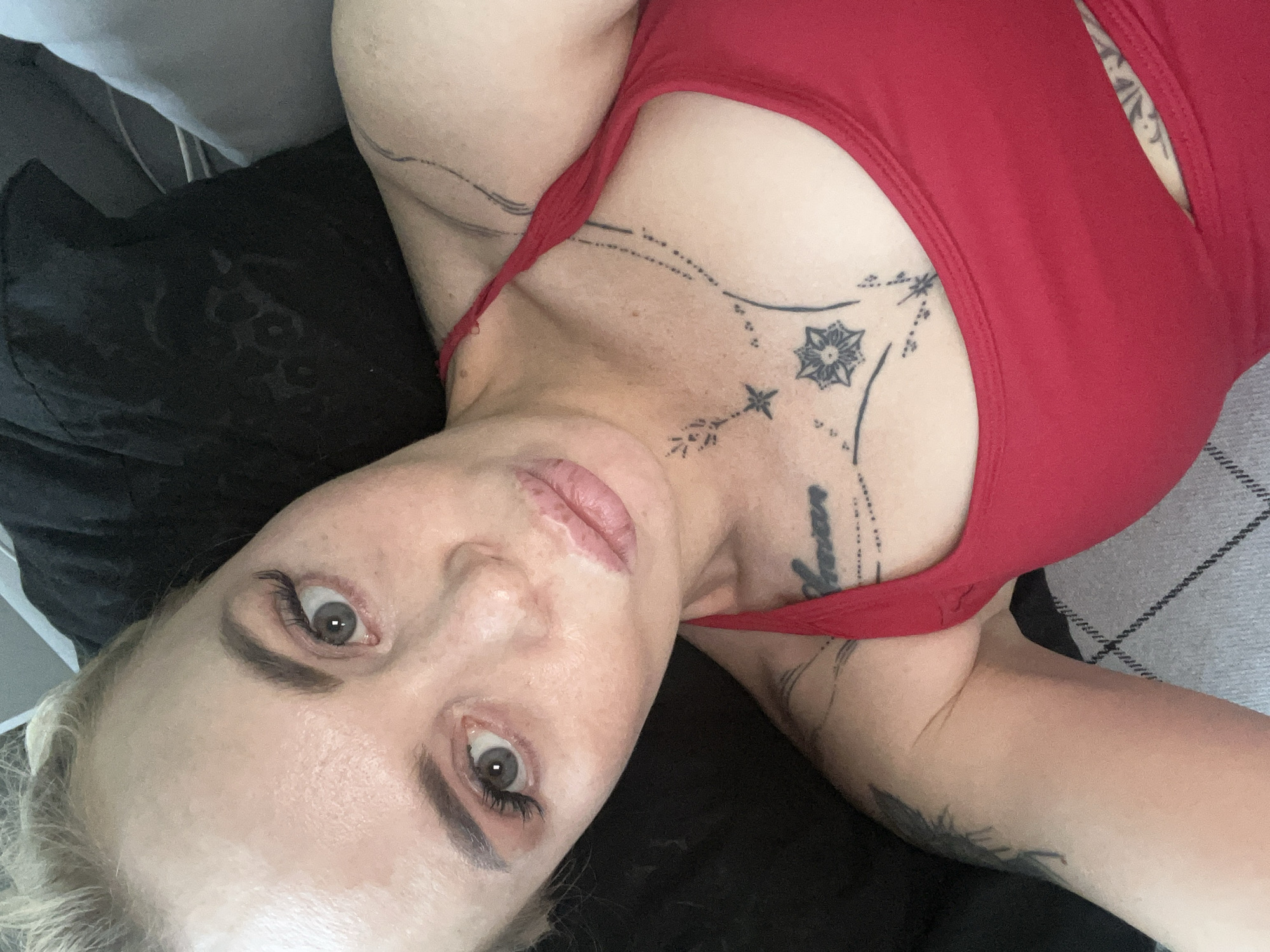 A woman with blonde hair and light-colored eyes is shown in a close-up, overhead shot. She is wearing a red tank top and has several tattoos on her chest and arm. Her lips are slightly parted, and her eyes are looking towards the camera. The background consists of dark pillows and a gray and white patterned fabric. The scene appears to be indoors.