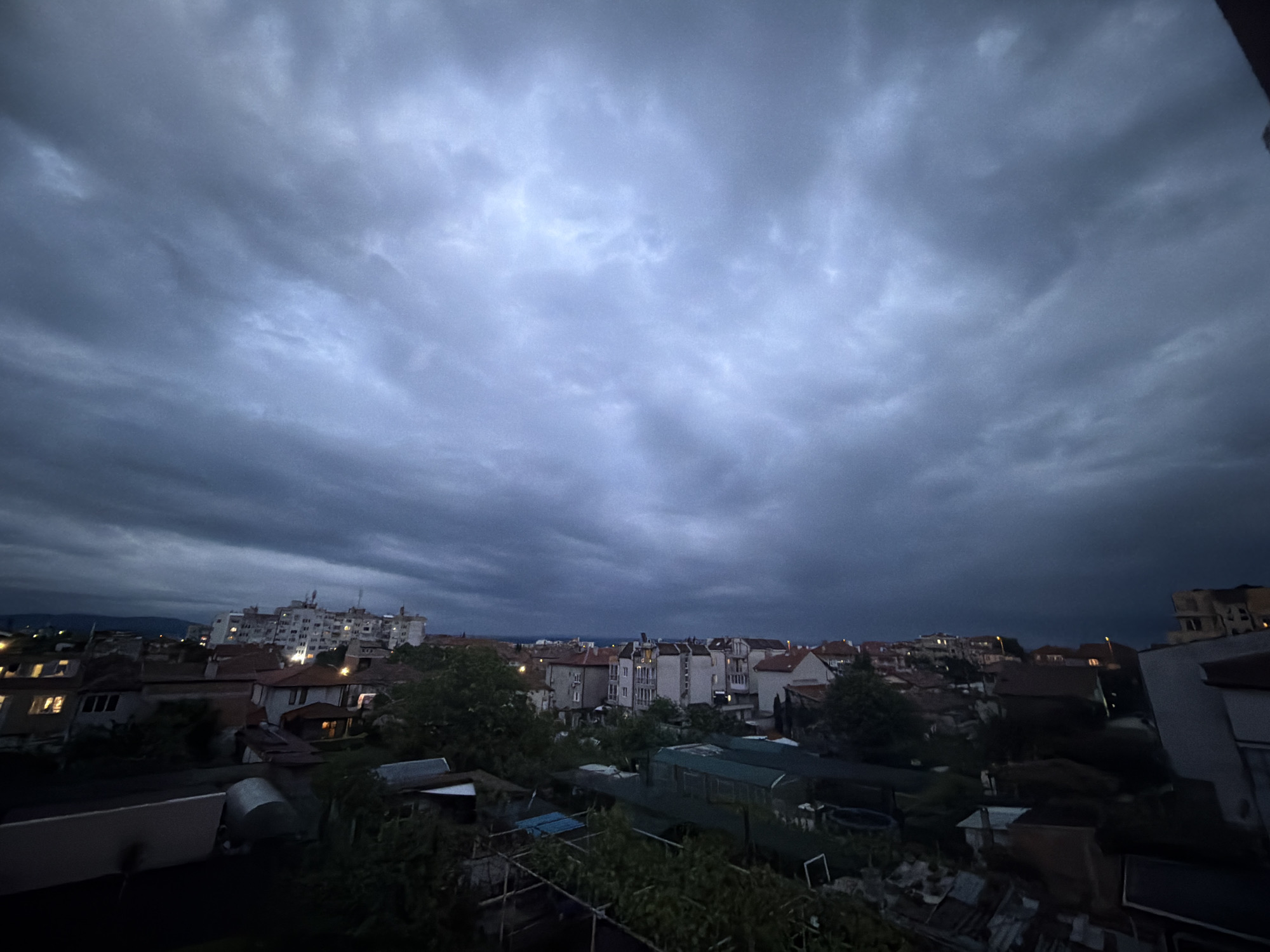 The image depicts an overcast urban landscape, likely taken from an elevated vantage point, possibly a balcony or rooftop. The sky is filled with dark, ominous clouds, suggesting an approaching storm or recent rainfall. Below, a dense cluster of buildings with red-tiled roofs dominates the midground. Some buildings are multi-story apartment blocks, while others appear to be smaller residential houses. There are various outbuildings, sheds, and greenhouses visible. A few trees provide some greenery. In the distance, a row of taller buildings is visible. The city is Burgas, Bulgaria.