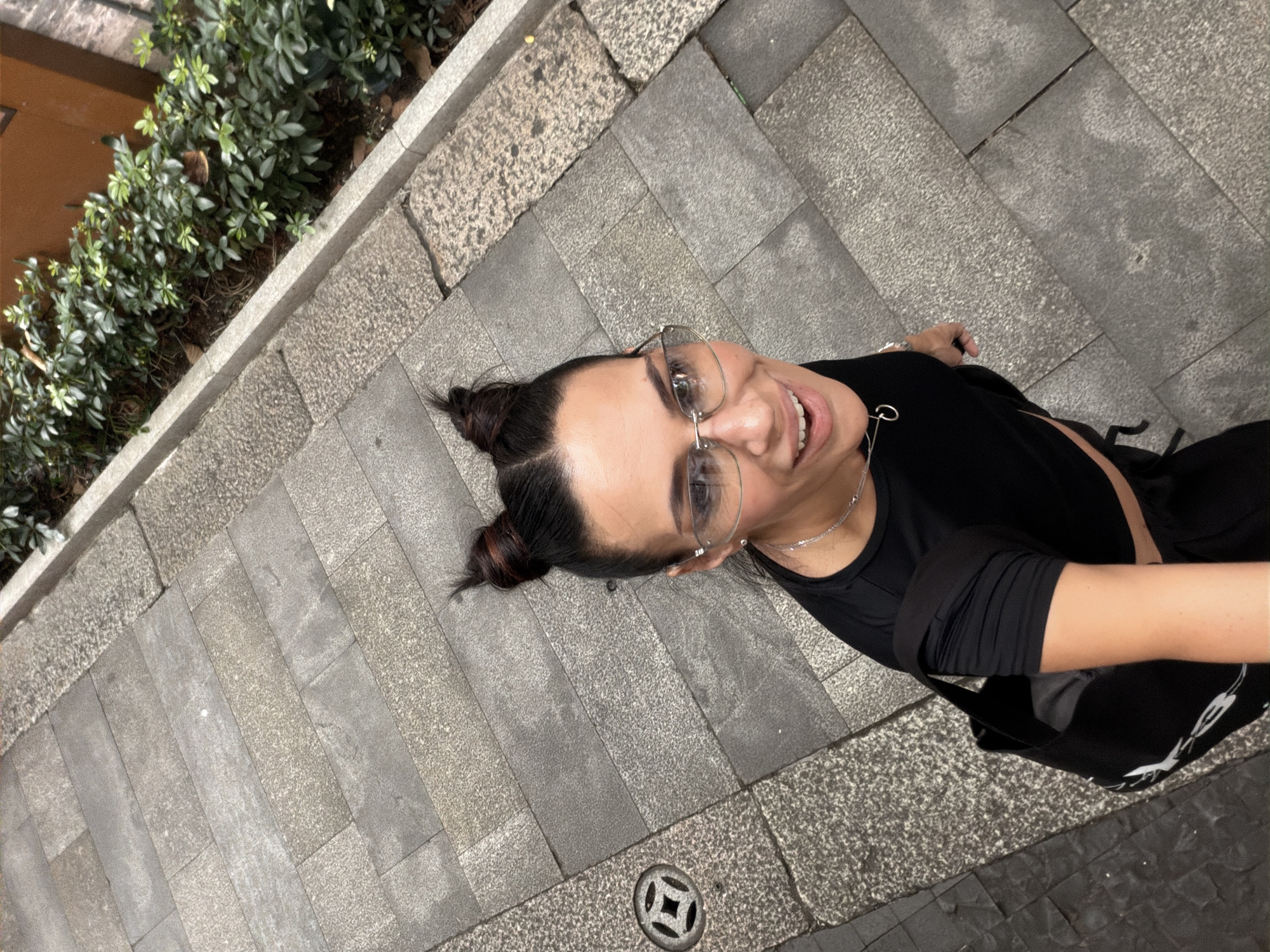 A high-angle, medium shot captures a smiling woman from the chest up, walking down stone steps. She has dark hair styled in two small buns on top of her head and is wearing clear-rimmed glasses, a black crop top, and a silver necklace. A black bag with a white logo is slung over her left shoulder. The background consists of stone pavement with various patterns and a line of green bushes on the right. A circular drain cover with a geometric design is visible on the left side of the steps.