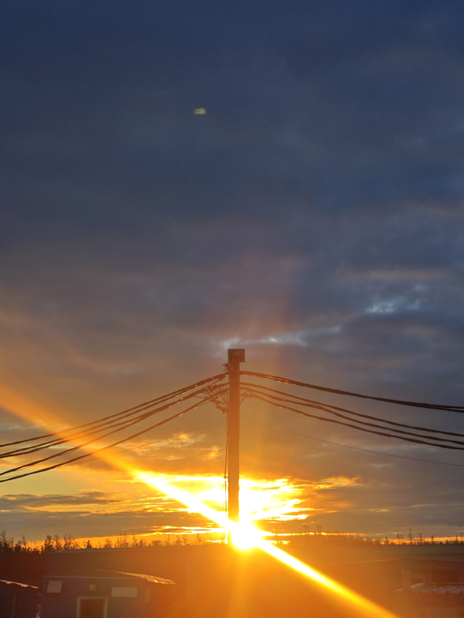 The image depicts a sunset in an unidentified location in Russia. The scene is dominated by a bright, orange sun setting behind a utility pole with several power lines extending from it. The sun's rays create a strong glare. The sky is a mix of dark blue, gray, and orange clouds, with a possible faint celestial object visible in the upper portion of the image. The silhouette of trees is visible along the horizon. In the foreground, a dark, rectangular structure resembling a small building is partially visible.