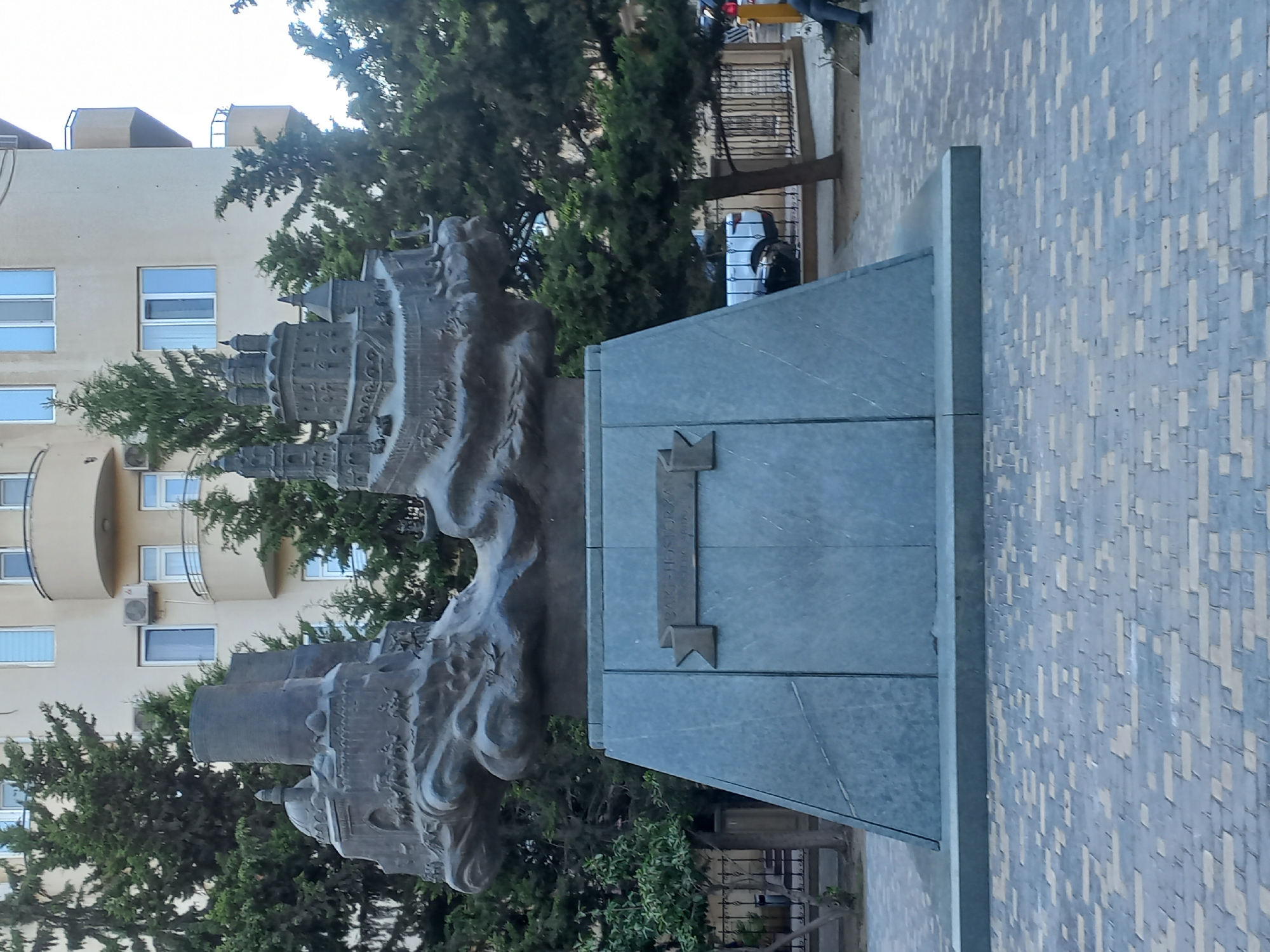 A sculpture depicting a historical scene or city is displayed on a stone pedestal in a public square in Baku, Azerbaijan. The sculpture, made of dark metal, features architectural elements like towers, domes, and fortifications, along with small figures that appear to represent people or animals engaged in some activity. The pedestal is pyramidal and made of a light gray, possibly granite, material. A banner-like plaque is affixed to the front of the pedestal, bearing an inscription. The sculpture is situated in front of a modern apartment building with multiple balconies and windows. Evergreen trees are visible behind and around the sculpture, and the ground in the foreground is paved with interlocking bricks in shades of gray and beige. A metal fence runs along the background. A white car and a person in dark clothing sitting on the ground are visible to the right of the sculpture. The overall scene appears to be an outdoor public space in a city.