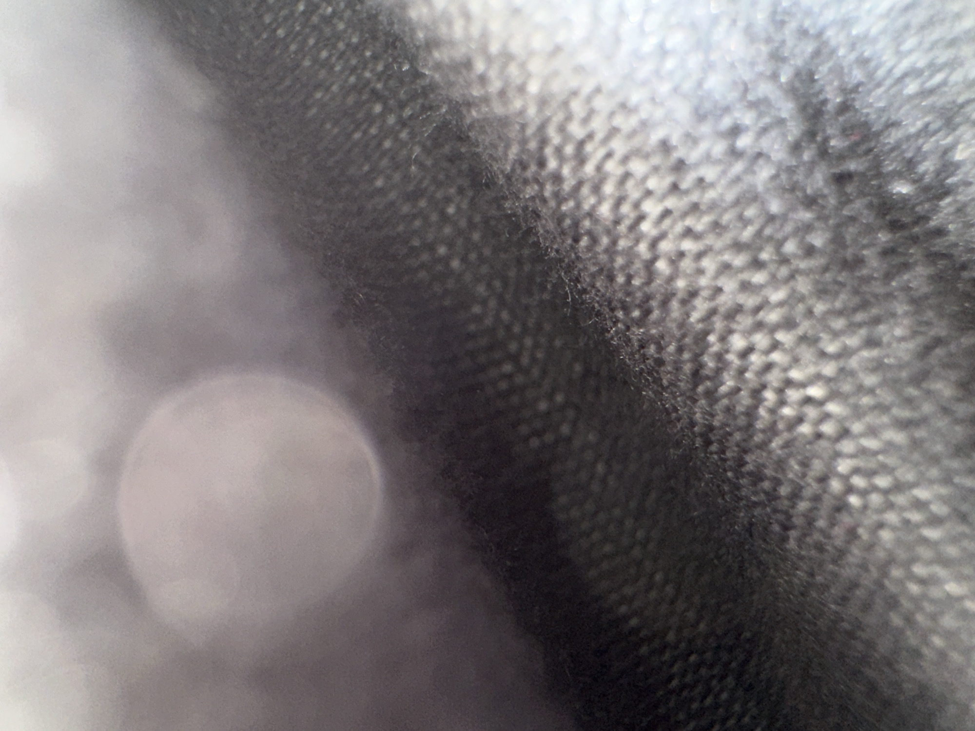 The image is an extreme close-up of a dark, textured fabric, likely a coarse weave like denim or canvas. The fabric is angled, with a darker area on the left transitioning to a lighter, more reflective area on the right. There is a large, out-of-focus circular light source or reflection in the upper left quadrant of the image, casting a soft glow. The texture of the fabric is emphasized, showing individual threads and the pattern of the weave. The image appears to be taken with a shallow depth of field, rendering much of the scene out of focus.