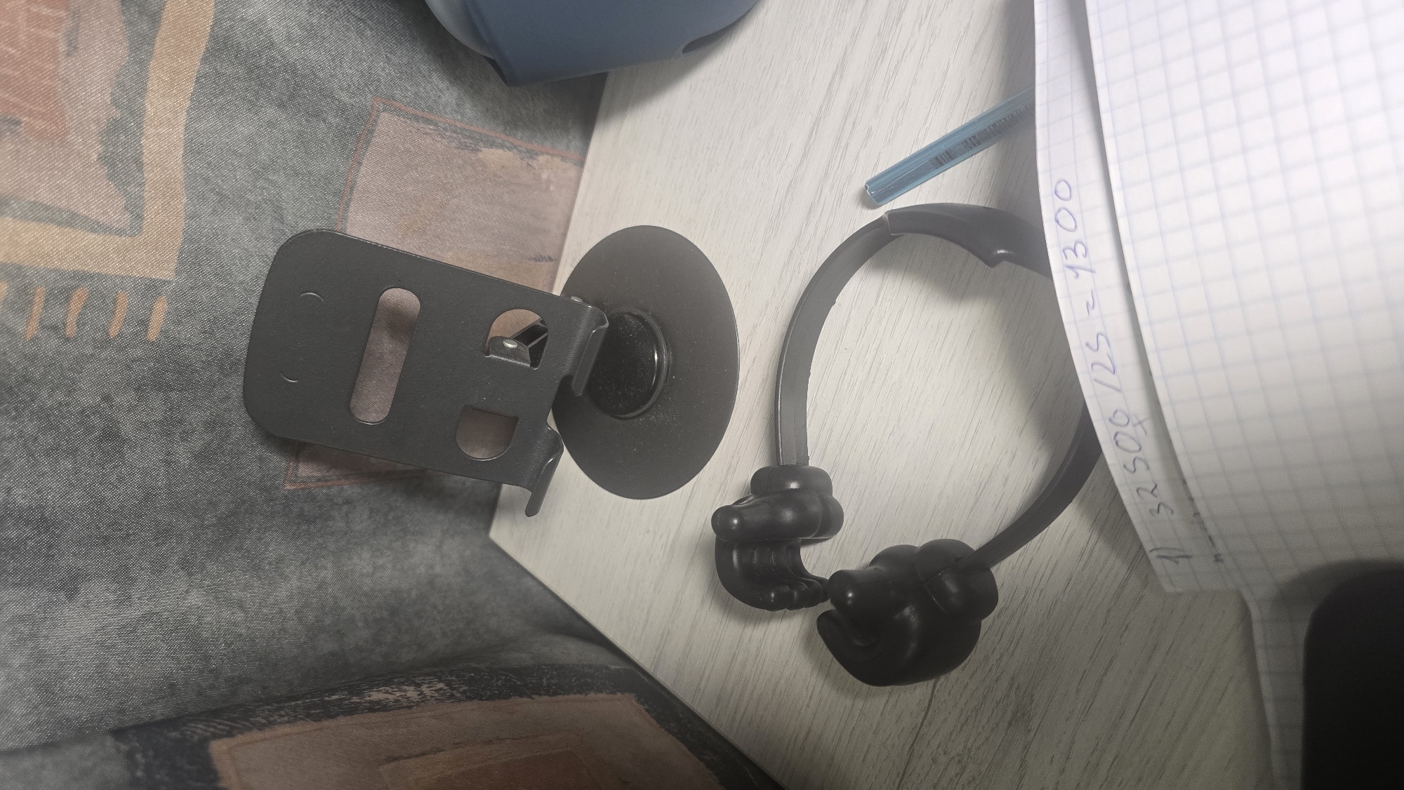 The image shows a close-up, slightly overhead view of a desk with a notebook, a phone stand, and a pair of novelty headphones. The notebook is open to a page with blue graph paper, and a handwritten mathematical equation is visible: "1) 32500/25 = 1300". The phone stand is black, with a circular base and a vertical arm that holds a phone in place. The headphones are black and designed to look like hands giving a thumbs-up. A blue pen rests on the notebook. The background is a textured fabric surface, possibly a sofa or curtain. The lighting is artificial and casts shadows. There is no discernible indication of the location in Ukhta, Russia.