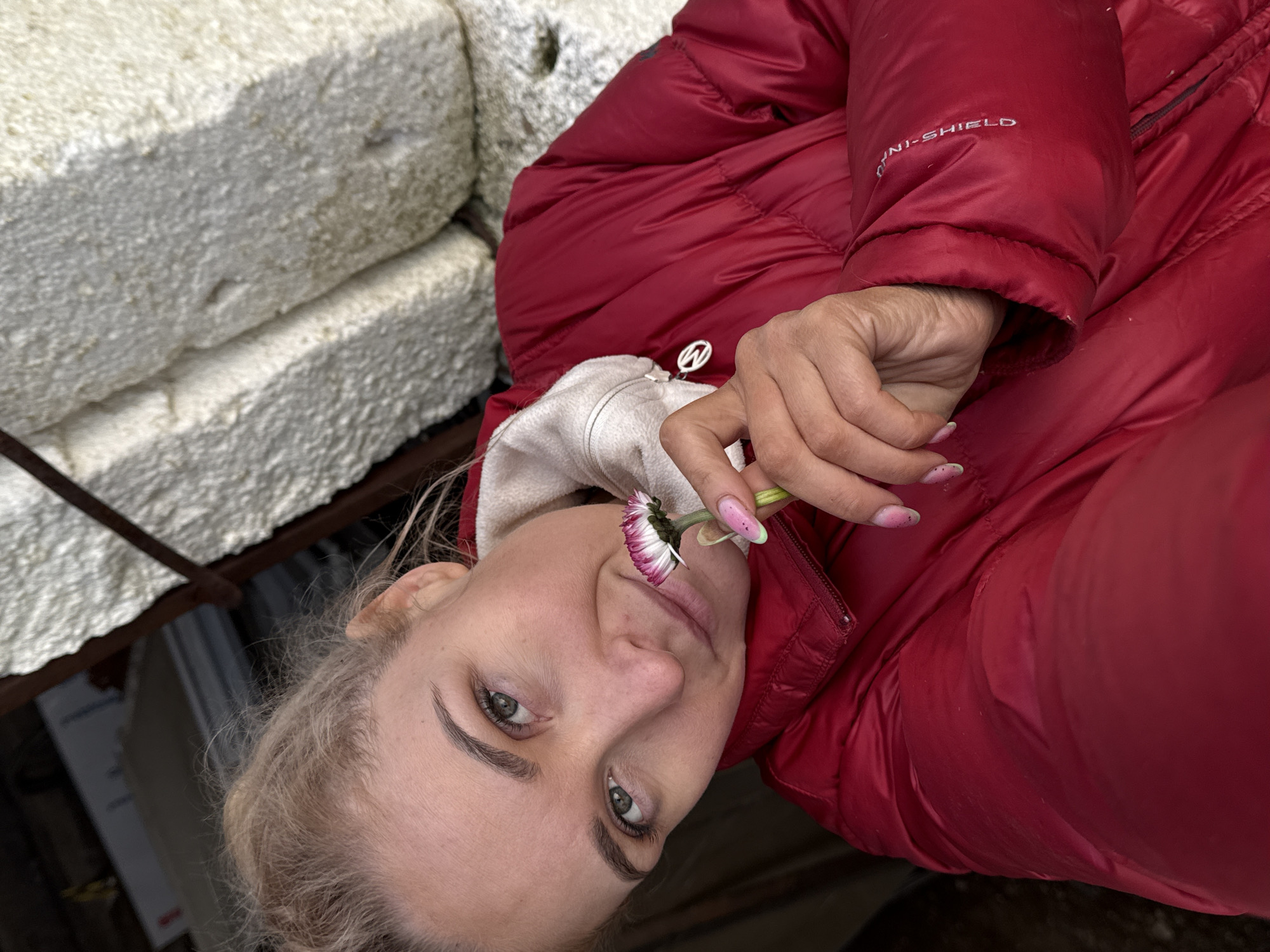 A woman with blonde hair holds a flower near her face. She is wearing a red, puffy jacket with "OMNI-SHIELD" visible on the sleeve. In the background are light-colored, rectangular blocks. The location in Rosstani, Russia, is not discernible from the given image.