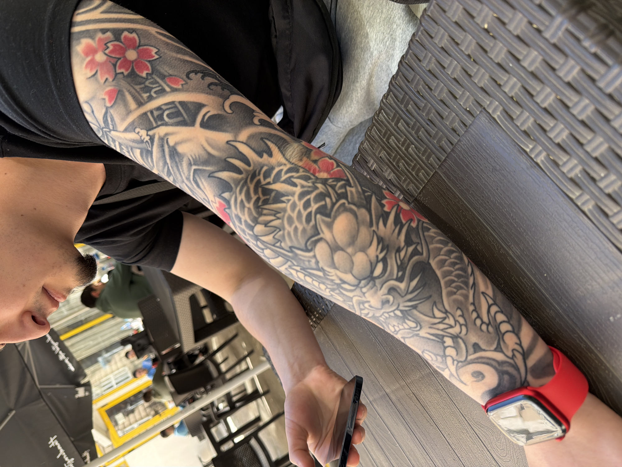 A close-up, high-angle shot captures a person's left arm, heavily tattooed, and their partially visible face and torso in an indoor setting in Plovdiv, Bulgaria. The individual, wearing a black t-shirt, has a full-sleeve Japanese-style tattoo covering their entire arm. The intricate design features a large, detailed dragon with scales and waves, predominantly rendered in shades of black, gray, and white. Red cherry blossoms add vibrant splashes of color, primarily near the shoulder and upper arm.
The person holds a dark-colored smartphone in their left hand, while a red-banded smartwatch with a clear, reflective screen is visible on their right wrist. Their face, seen from the chin up, shows a light mustache and stubble. In the blurry background, other individuals are present, along with what appears to be an industrial or commercial interior. Yellow lines are painted on the floor, and metallic structures are visible. A black studio light stand, partially obscured, has faint, partially legible text "fomny" on it, suggesting a creative space or studio environment.
The person is seated at a table or desk, evidenced by the textured, dark wicker-patterned surface beside their arm and a darker wooden floor section. The overall lighting is bright, indicating daytime or a well-lit indoor area. The scene appears casual and relaxed, with the person likely browsing or interacting with their phone.
