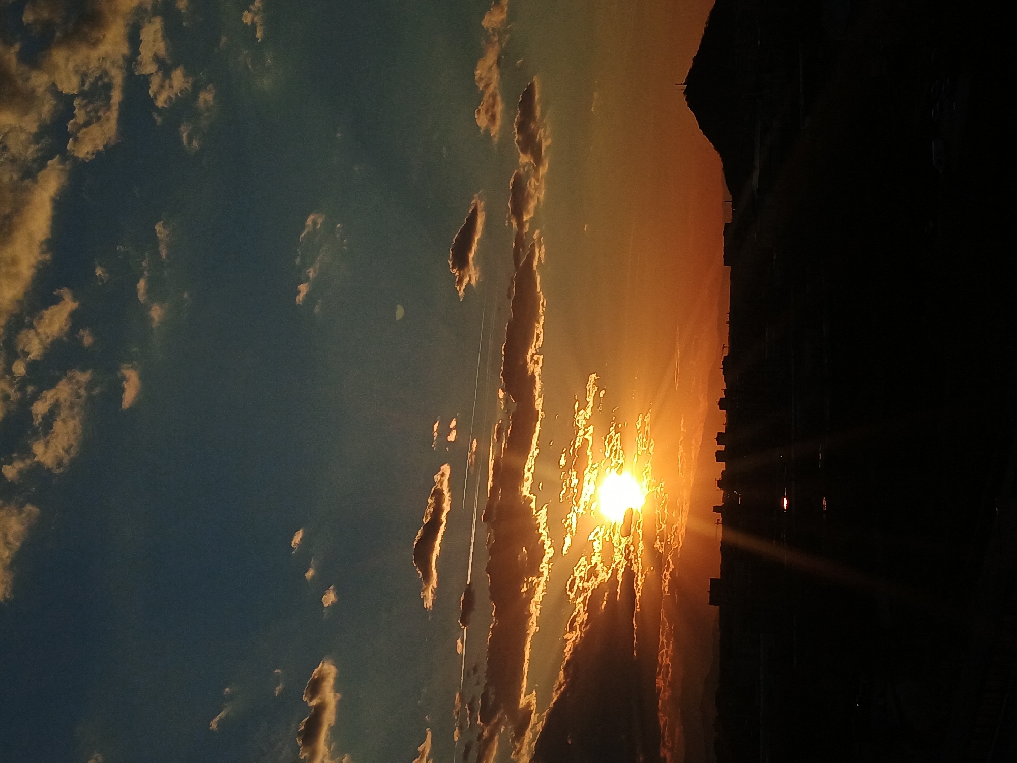 The image depicts a sunset in Plovdiv, Bulgaria, with the sun low on the horizon, obscured by and shining through clouds. The sky transitions from a deep blue to a bright orange near the sun. A dark silhouette of buildings is visible against the bright sunset on the right side of the image. There are contrails in the sky, likely from a passing airplane. The image is oriented sideways.
