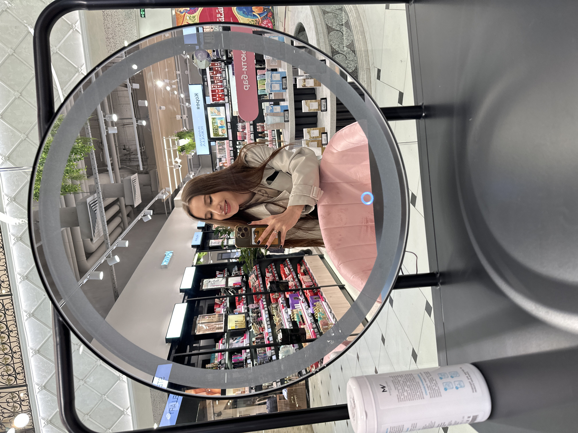 A young woman is captured taking a selfie in a large, round, illuminated mirror within a retail store or shopping mall. The image is taken at an angle, causing the reflection in the mirror to appear rotated.
The woman, with long brown hair, is wearing a light-colored jacket and is smiling while holding her smartphone, which she uses to photograph her reflection. She is seated in a pink upholstered chair. The mirror features a built-in ring light and a glowing blue power button on its right side.
The reflection in the mirror reveals the store's interior, characterized by numerous shelves fully stocked with products, likely cosmetics or beauty supplies. The ceiling, also visible in the reflection, features modern lighting, air vents, and decorative panels, including an ornate section with a patterned grid. The floor visible in the reflection and outside the mirror's frame is tiled in a black and white checkerboard pattern.
In the foreground, outside the mirror's reflection, a white cylindrical bottle with a blue label and an "MV" logo is placed on a black surface. Reflected text observed in the store background includes "Конеё" (likely a mirrored "Конец" meaning "End" in Russian), a partial "PRO-N-dp", "HERVAGH" (possibly a mirrored brand name), and "BALET". The setting is brightly lit, indicative of a commercial environment, and the woman appears cheerful.