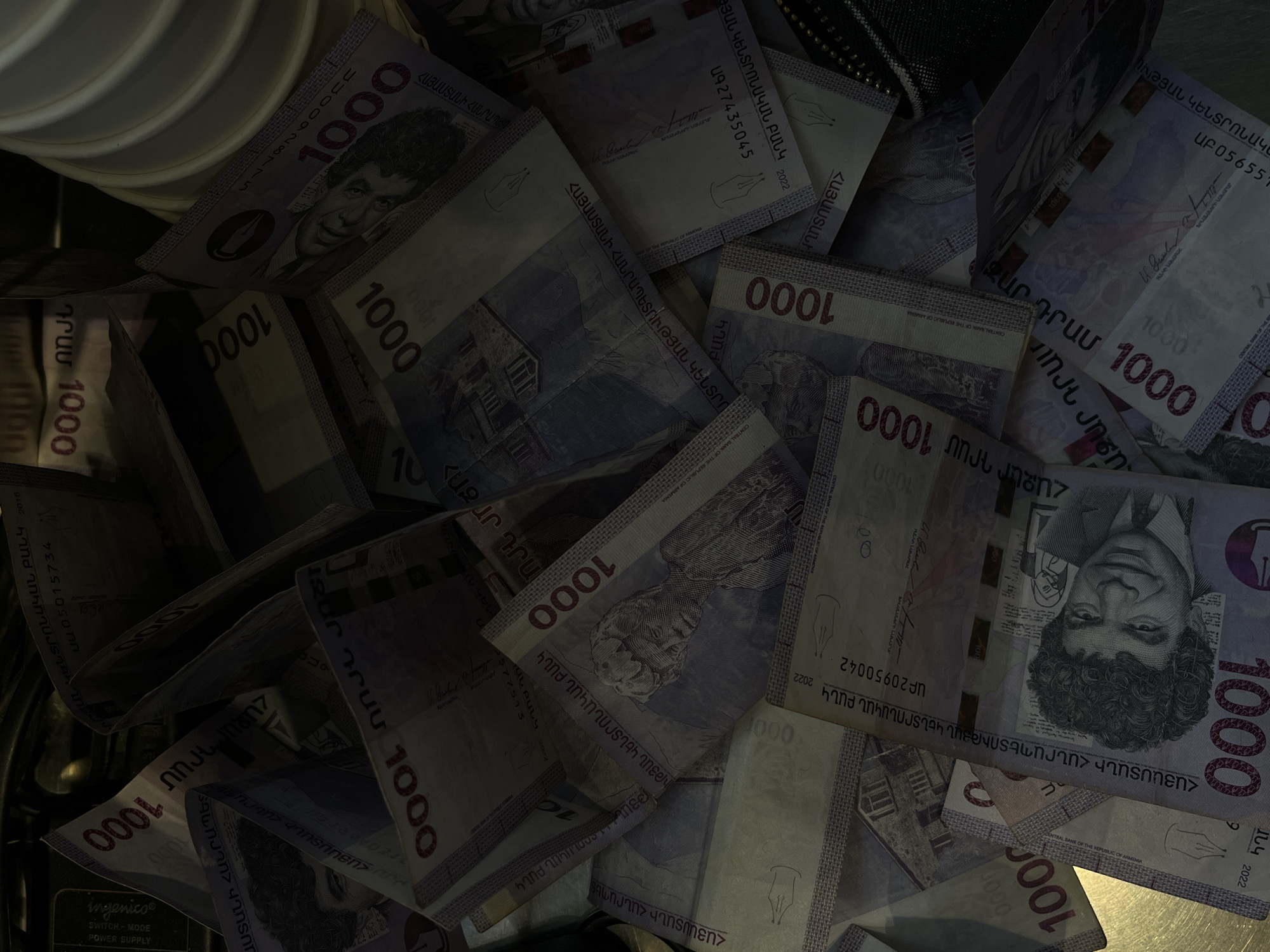 The image shows a close-up, overhead view of a collection of Armenian 1000 dram banknotes. The banknotes feature a portrait of Paruyr Sevak and are dated 2022. The notes are scattered and partially overlapping. The composition is densely packed, with the banknotes filling most of the frame. Some of the notes show their serial numbers and signatures. The background is dark and obscured, and the scene is possibly contained within a drawer or container. In the top left of the image, there is a stack of what appear to be white ceramic or plastic items.