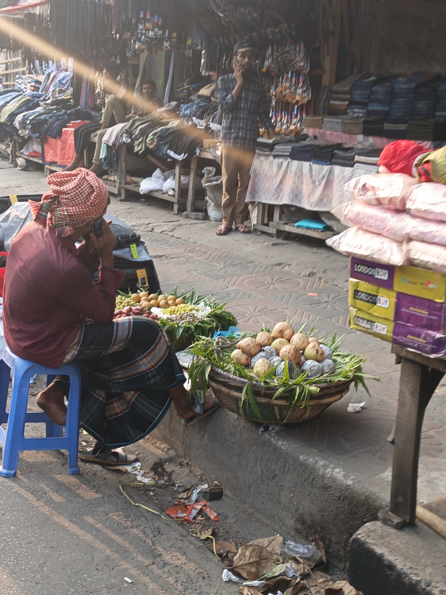 This image depicts a vibrant and somewhat cluttered street market scene in Dhaka, Bangladesh, illuminated by strong sunlight that creates a prominent lens flare and long shadows, suggesting either morning or late afternoon.
In the foreground on the left, a man is seated on a blue plastic stool, his back to the viewer. He wears a maroon long-sleeved shirt, a dark, striped lungi, and a red and white checkered head covering, and is talking on a mobile phone. His bare feet are visible, and sandals rest on the ground beside him. Near his feet, the ground is littered with dry leaves, plastic, and other debris. Adjacent to him, two large, round woven baskets overflow with fresh produce: one contains numerous light-colored, round fruits (possibly guavas) nestled among green leaves, and the other holds smaller yellow/orange fruits and red onions.
To the right, stacked cardboard boxes are visible, prominently displaying the text "LondonBoy" in yellow and purple. Further into the midground, a man wearing a plaid shirt, light-colored pants, and a cap stands near a market stall, also talking on a mobile phone. He appears to be a vendor. The market stalls lining the background are constructed from wood and display a variety of merchandise, including folded clothes (shirts, pants) and various accessories hanging up. Several other individuals can be seen in the background, sitting or standing around the stalls, contributing to the busy atmosphere of daily commerce.
The scene captures the informal yet bustling nature of a typical South Asian street market, characterized by close quarters, diverse goods, and active social and commercial exchanges.