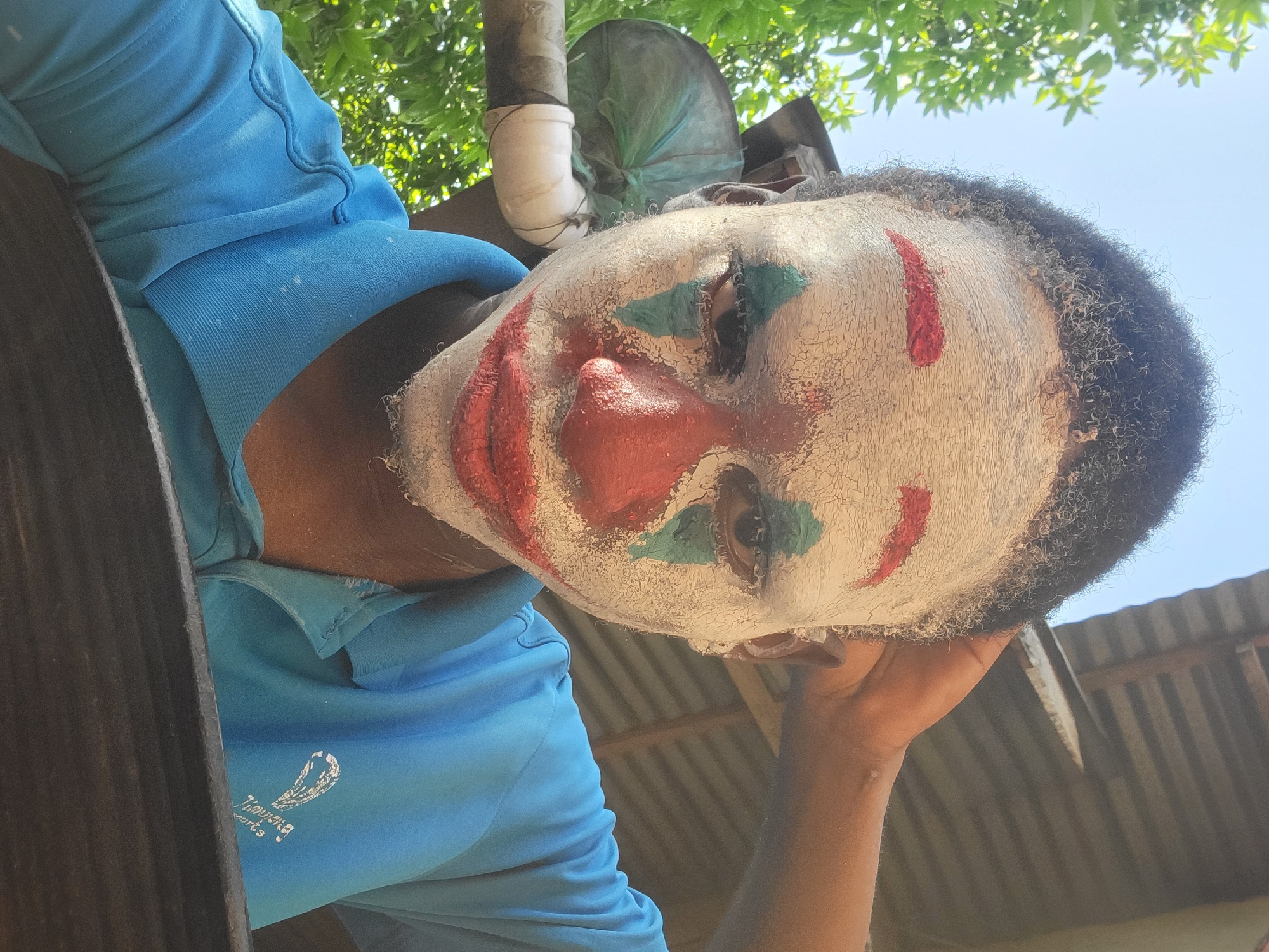 A person wearing clown makeup and a blue shirt stands outside in Nkpor, Nigeria. The makeup includes a white face with cracked texture, red lips and nose, and red and green markings around the eyes. Above, there is foliage and a piece of plumbing. The person is looking down at the camera. Corrugated metal roofing is visible in the background. The logo on the person's shirt reads "Flourishing Resorts."