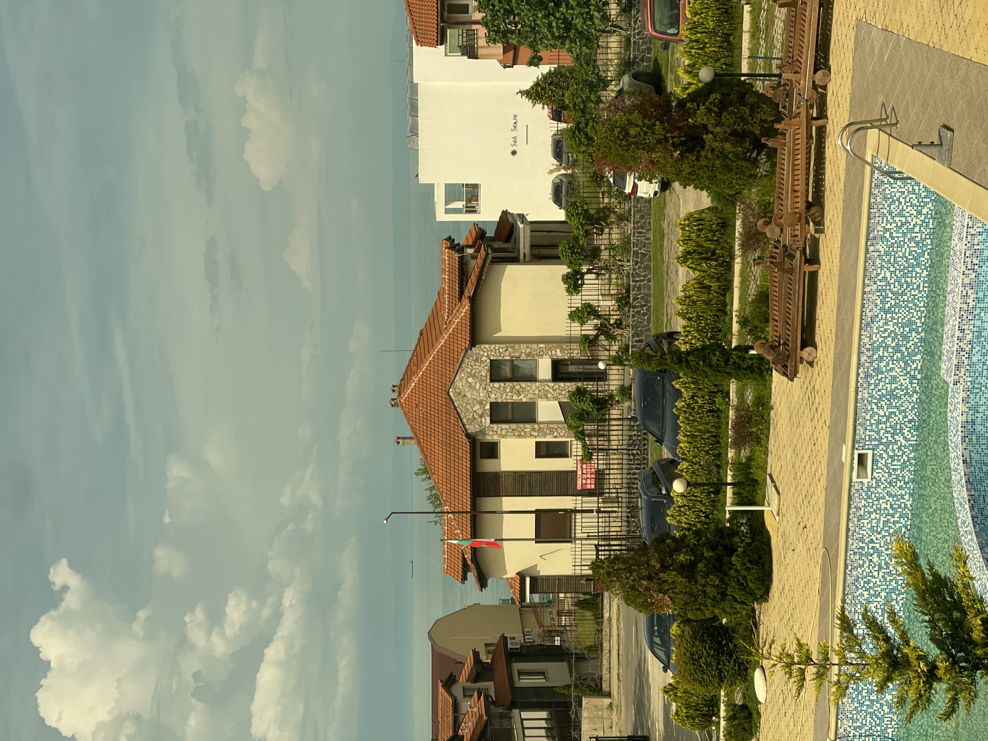 The image shows a street scene in Sozopol, Bulgaria. The street is lined with multi-story buildings with red-tiled roofs. A Bulgarian flag is visible in front of one of the buildings. There are parked cars along the street. The sky is cloudy. A swimming pool is visible on the right side of the image.