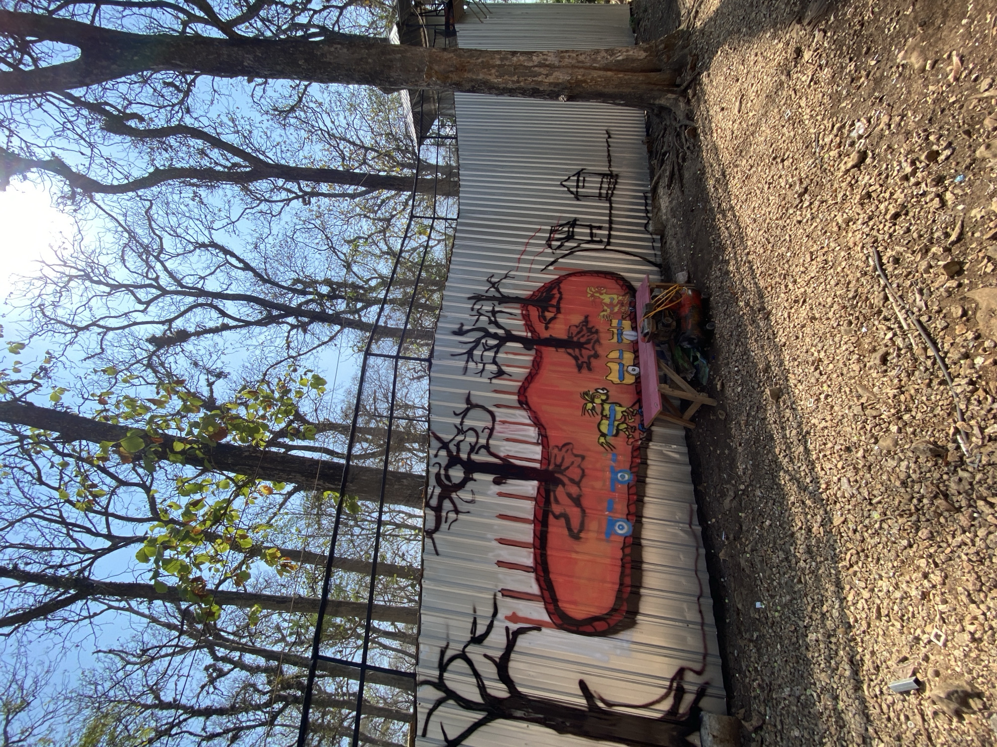 The image shows a corrugated metal fence with graffiti on it. The graffiti depicts a large orange oval with black outlines of trees and what appear to be cartoon characters driving small vehicles within it. To the left of the oval, there is a black silhouette of a bare tree. To the right of the oval, there are simple black outlines of two houses or structures. In front of the fence, there is a small wooden bench painted pink, with some equipment like a red compressor and yellow hoses underneath it. The ground in front of the fence is covered with small stones and dirt, with some scattered debris. Behind the fence, there are many tall trees with bare branches and a clear blue sky. The overall scene appears to be outdoors, possibly in a park or a less developed area. There are no people visible in the image. The location is described as Unknown, Unknown.