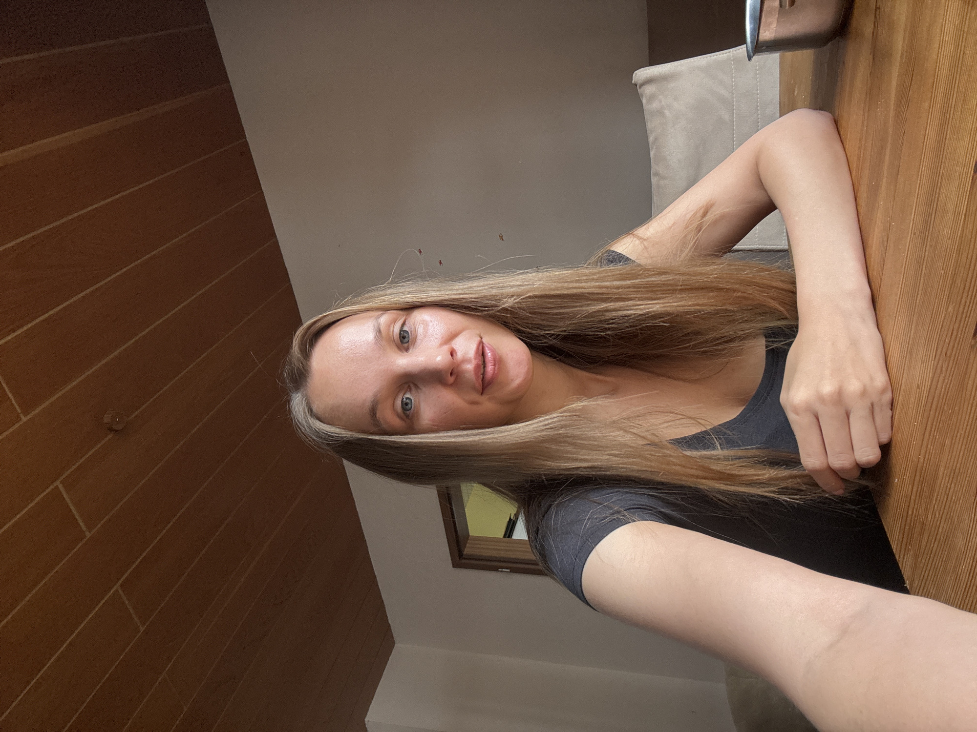A young Caucasian woman with long blonde hair and blue eyes sits at a wooden table. She is wearing a dark grey t-shirt. Her left arm is extended towards the camera, and her right arm is resting on the table. Behind her, a beige chair is visible, and to the far right, a metal bowl sits on the table. The room has a wood-paneled ceiling and a light-colored wall. A door with a yellow interior is visible in the background to the left. The scene appears to be an indoor setting, likely a home, with natural light illuminating the woman. The image was taken in Überherrn, Germany.