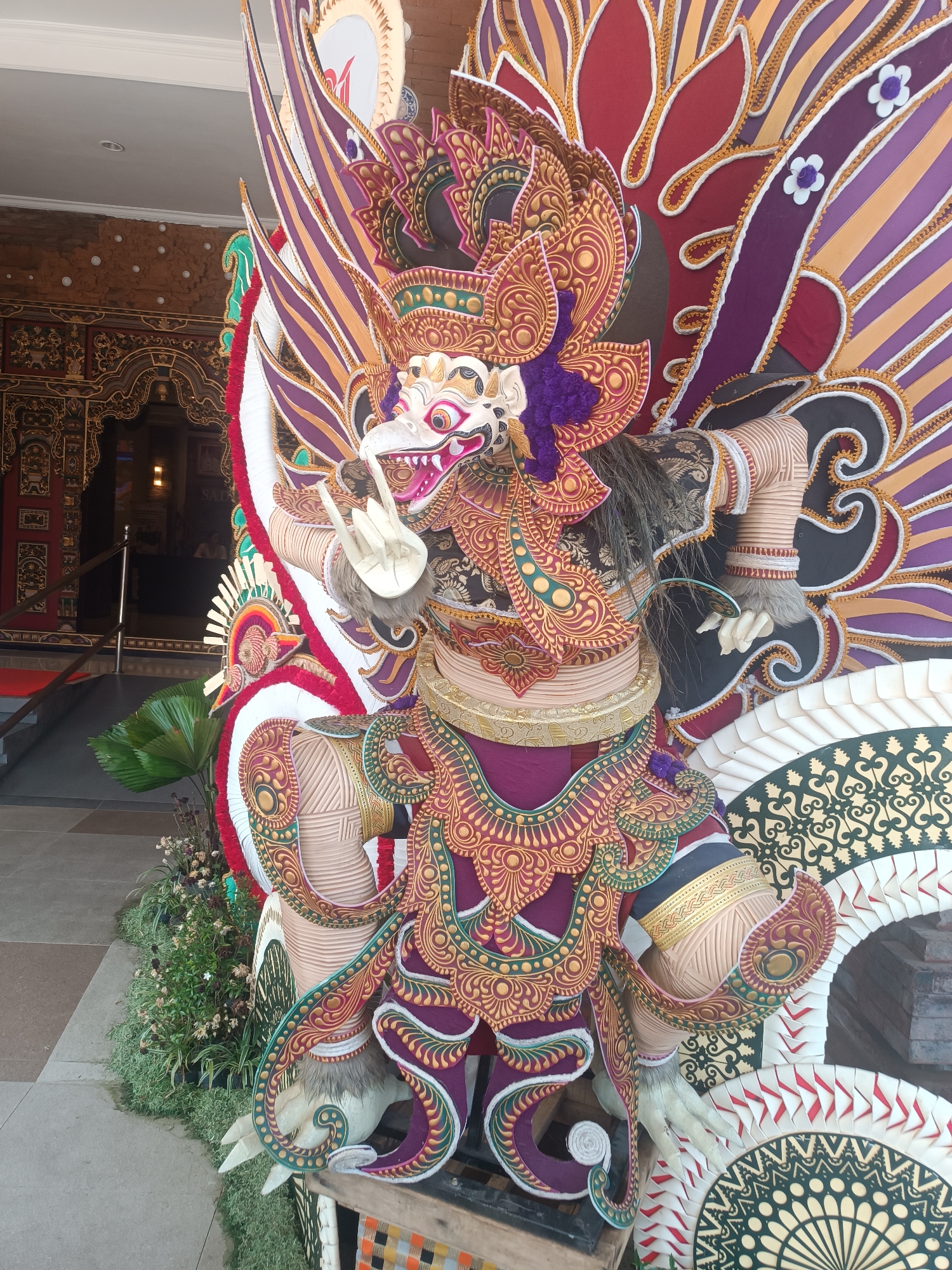 A large, colorful statue of a mythological creature, likely a Garuda, stands prominently in front of an ornate building entrance. The statue is elaborately decorated with intricate patterns and vibrant colors, including shades of purple, red, gold, and green. It features a bird-like head with sharp teeth and a fierce expression, large feathered wings, and human-like arms and legs adorned with traditional attire. The statue is positioned on a wooden platform, surrounded by lush green foliage and decorative elements. In the background, an arched entryway with intricate carvings and red doors suggests a cultural or ceremonial setting. The overall scene depicts a striking example of Indonesian artistry, possibly for a festival or religious event, located in Tabanan, Indonesia.