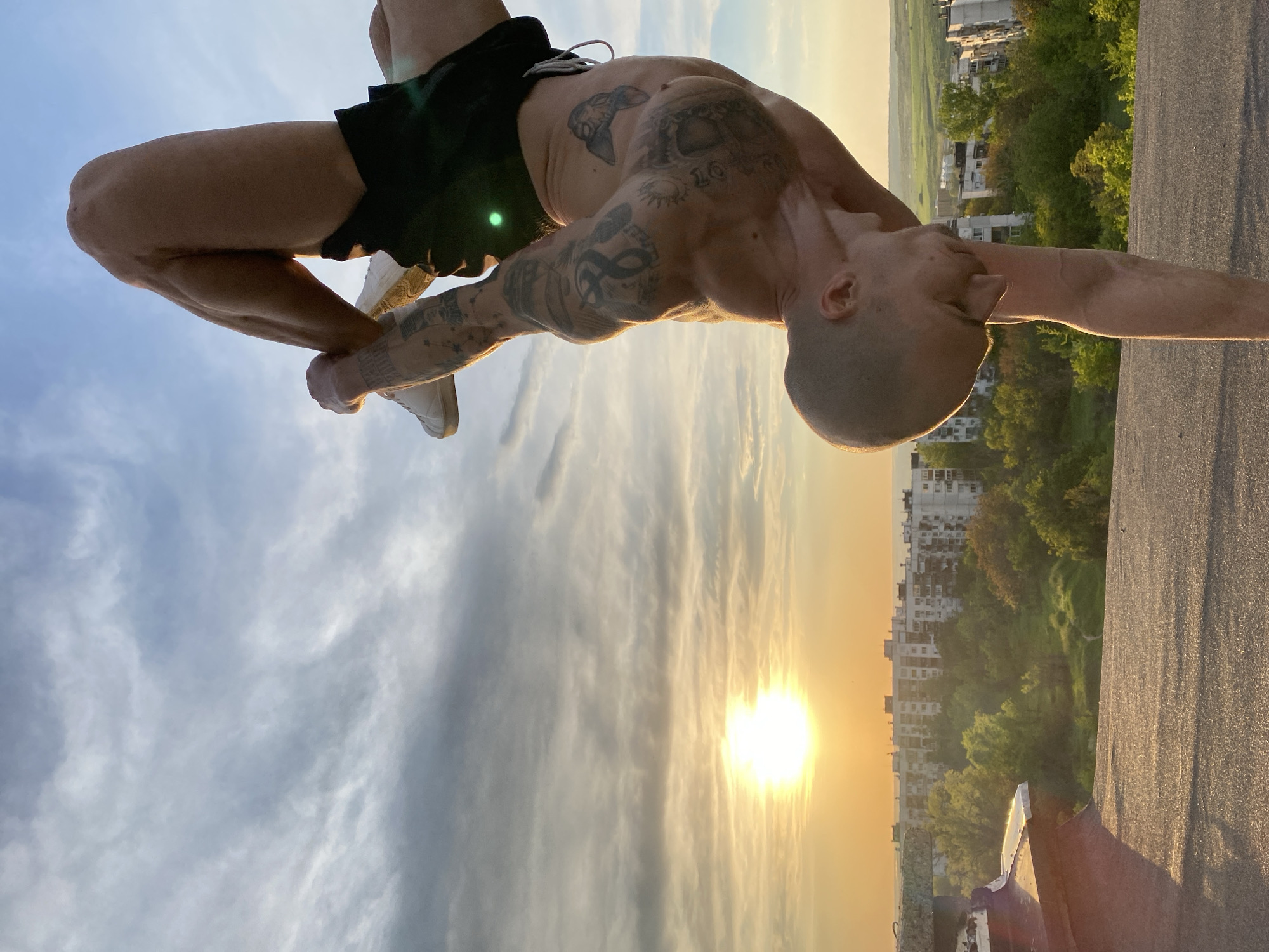 A tattooed man balances upside down on the edge of a building. He is wearing black shorts and white sneakers. The background shows a setting sun over a city landscape with buildings and trees. The man appears to be performing a stunt or calisthenics exercise. This appears to be a residential or urban area in Pleven, Bulgaria.