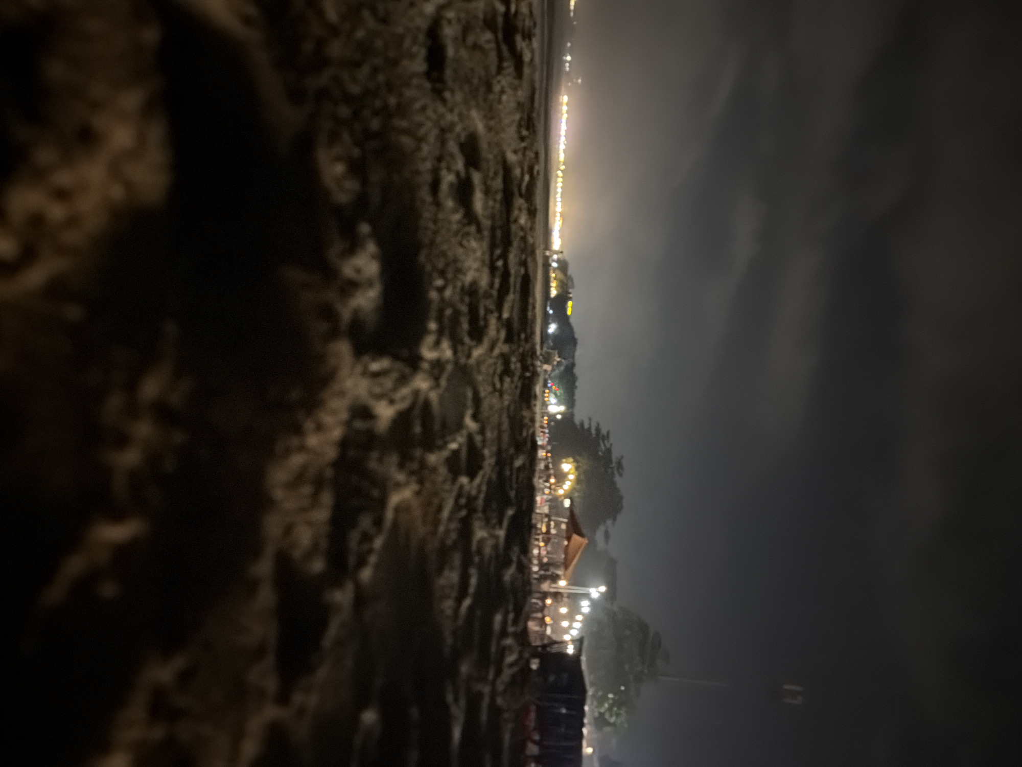 The image depicts a nighttime scene on a sandy beach in Kuta Selatan, Indonesia. In the foreground, out of focus, is the textured surface of wet sand. The midground and background reveal a lively beachside setting illuminated by numerous lights. Several small open-air restaurants or stalls are visible, with tables and chairs arranged under awnings. Strings of lights are hung between structures and along the shoreline, casting a warm glow. In the distance, a line of lights suggests a more populated area or the continuation of the beachside establishments. The sky above is dark with diffused clouds, and a tall pole with a light fixture is visible on the right side of the frame. No people are clearly discernible due to the low light and focus. The overall impression is of a vibrant night market or dining area on a beach.