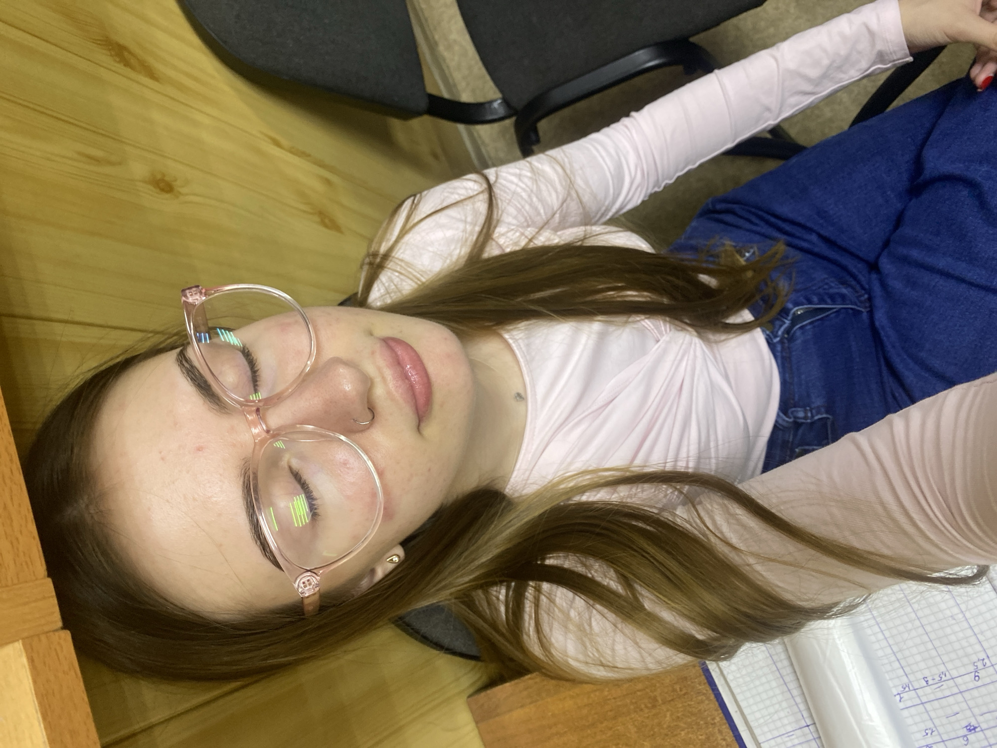 A young woman with long, brown hair and light skin is pictured from the chest up, seated in what appears to be a classroom or office setting. She is wearing large, translucent pink-rimmed glasses and a light pink long-sleeved shirt with a ruched neckline. Her eyes are closed, and she has a silver nose ring and a small silver necklace. Her lips are slightly parted, and she is wearing subtle makeup. Her skin shows some redness and visible pores. She is wearing dark blue jeans. To the left of the image, a portion of a wooden desk and a notebook with handwritten numbers and lines are visible. To the right, parts of two black office chairs are seen. The background consists of a wooden paneled wall. The overall lighting suggests an indoor environment. The location is identified as Dyakovo, Russia.