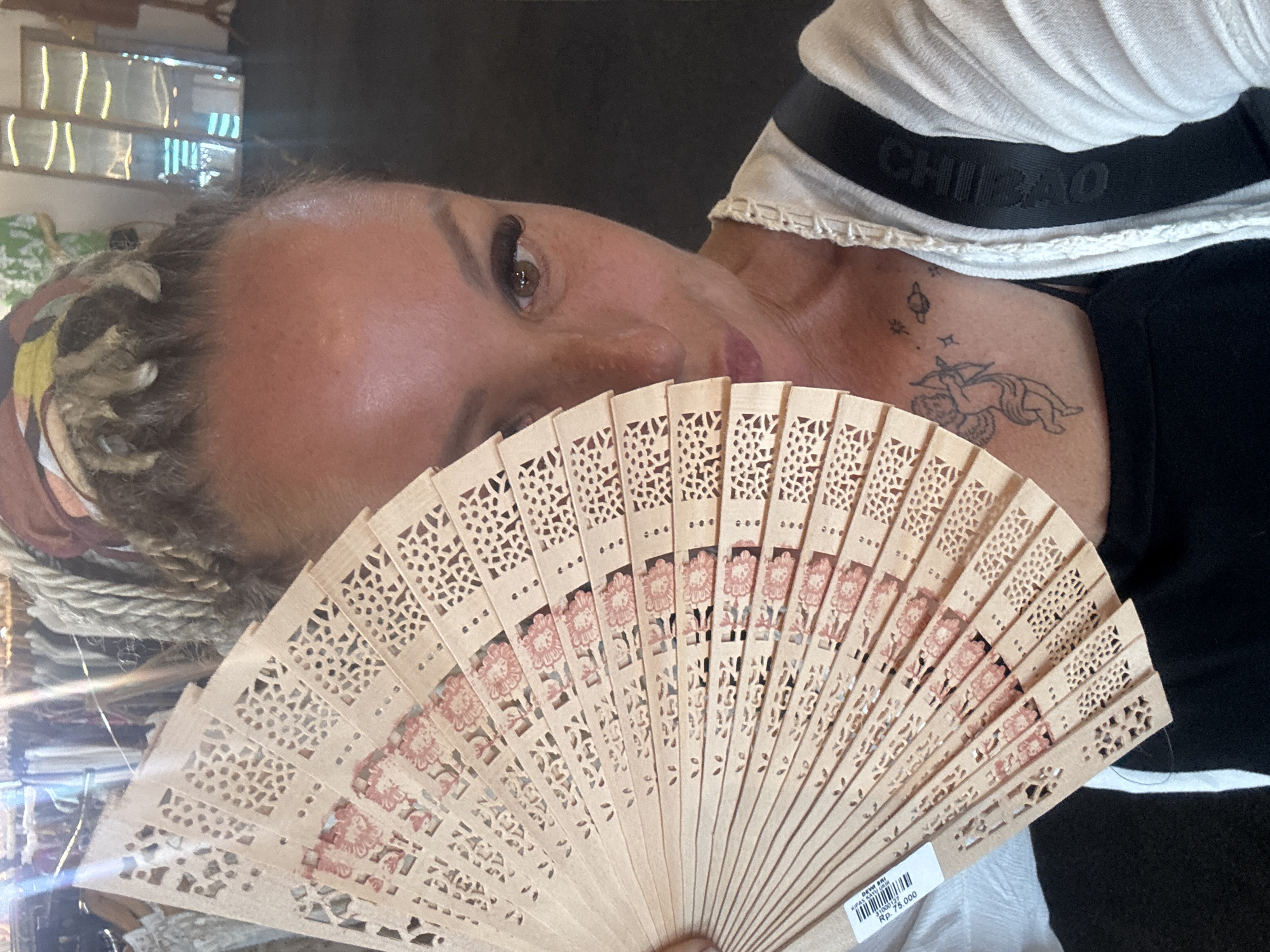 An indoor shot shows a person with braided blonde hair and a tattoo on their chest partially obscured by an open, tan, floral-patterned hand fan. The individual is wearing a black shirt, and a white shirt with black straps and the word "CHIBAO" on the strap is visible. The person is likely in the city of Kuta Selatan, Indonesia, given the location tag. A window with a reflective surface can be seen in the background.