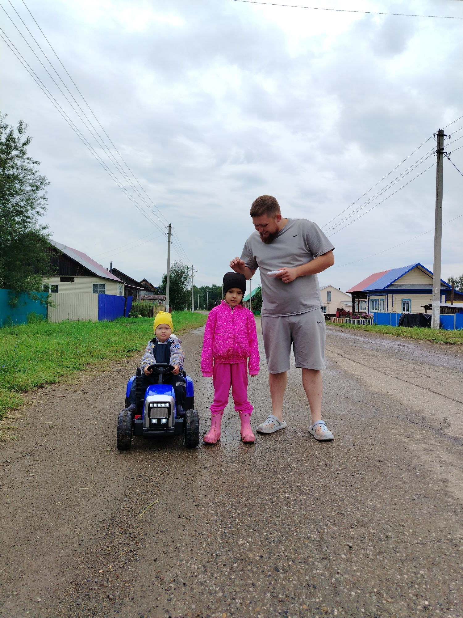 In Гожан, Russia, on an overcast day, a father stands on a gravel road next to his two children. The child on the left, wearing a yellow hat and a blue and grey jumpsuit, is seated in a blue toy tractor with the headlights on. To the right of the tractor, a young girl, dressed in a vibrant pink jacket and pants with pink boots, stands with her hands clasped in front of her. The father, clad in a grey t-shirt and shorts, is bent over slightly, touching the girl's head. In the background, houses with colorful roofs line the dirt road, and power lines stretch across the sky. The environment appears to be a rural residential area.