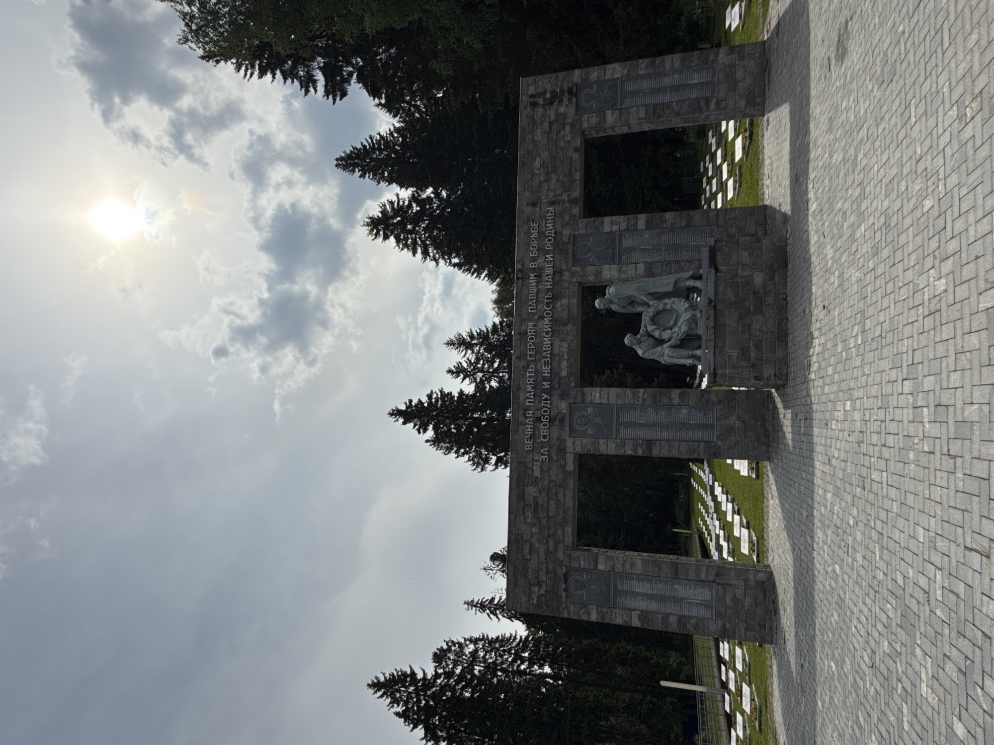 This image depicts a solemn war memorial or cemetery in Izhevsk, Russia, under a partly cloudy sky with bright sunlight. The central feature is a large, robust stone monument, constructed from rectangular block-like materials, forming an archway.
Within the archway, a dark-colored sculpture is prominently displayed, appearing to depict figures, one of whom is holding a wreath, conveying a sense of tribute and remembrance. Above this sculpture, on the horizontal beam of the monument, Cyrillic text is inscribed. It reads: "ВЕЧНАЯ ПАМЯТЬ ГЕРОЯМ, ПАВШИМ В БОРЬБЕ ЗА СВОБОДУ И НЕЗАВИСИМОСТЬ НАШЕЙ РОДИНЫ," which translates to "ETERNAL MEMORY TO THE HEROES WHO FELL IN THE FIGHT FOR THE FREEDOM AND INDEPENDENCE OF OUR MOTHERLAND." On the vertical pillars of the monument, there are additional plaques or panels with Cyrillic text, likely listing names or significant events, though the details are not clearly legible.
Behind the stone monument, a dense line of tall, dark evergreen trees, resembling fir or spruce, frames the scene against the sky. To either side and beyond the monument, neat rows of numerous white grave markers are visible on well-maintained green grass, confirming the setting as a memorial cemetery or burial ground. The foreground consists of a wide paved area made of light grey, rectangular paver blocks, suggesting a pathway or plaza leading to the monument.
The sky is partially covered with white and grey clouds, and the sun shines intensely from the upper left, casting a bright light over the scene. The time of day appears to be late morning or early afternoon. The overall atmosphere is one of respectful commemoration and serene remembrance.